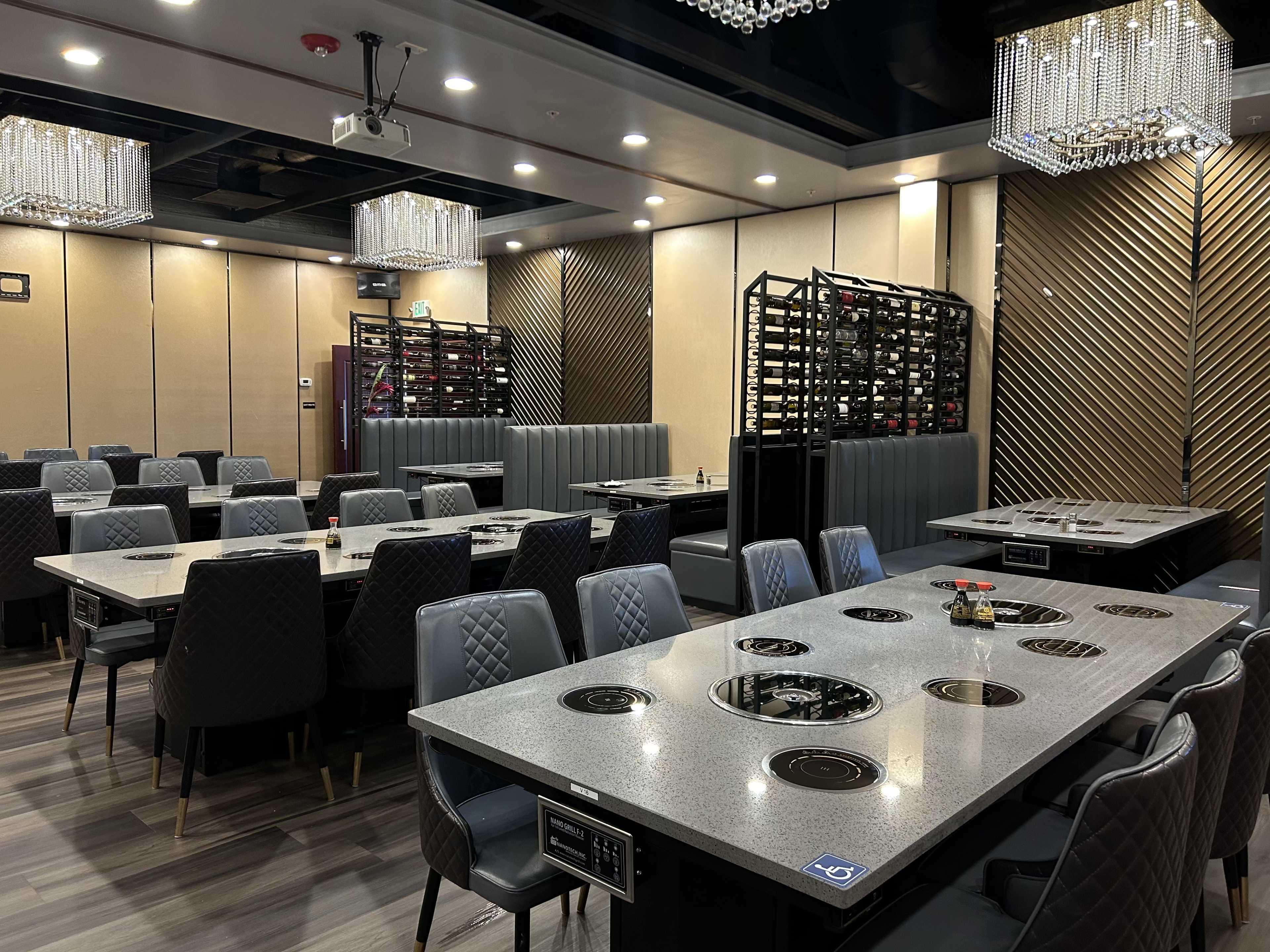 A modern restaurant interior with multiple dining tables featuring built-in cooking stations and a wall of wine racks in the background.