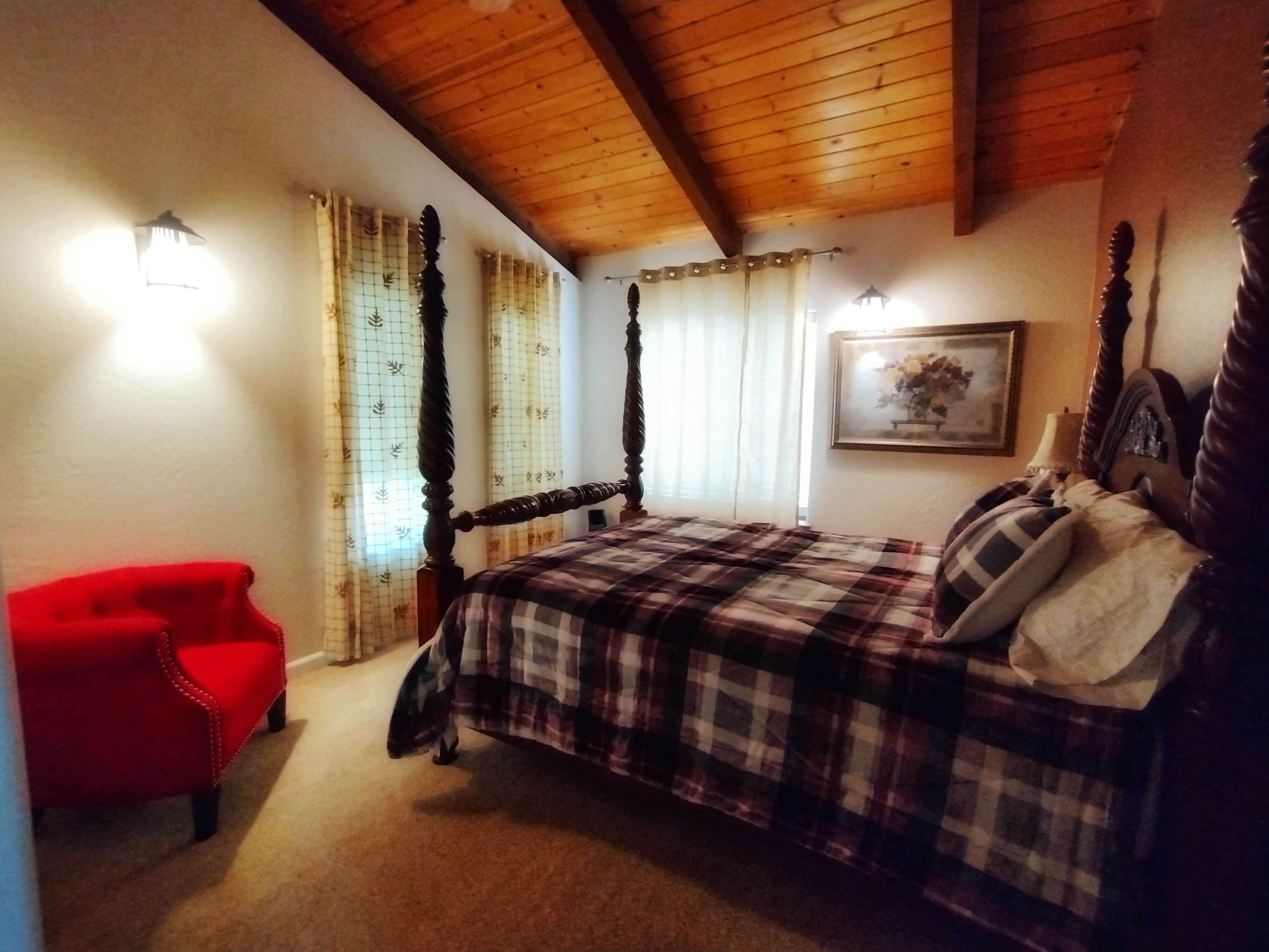 A bedroom with a four-poster bed, a red accent chair, and large windows dressed with light curtains.
