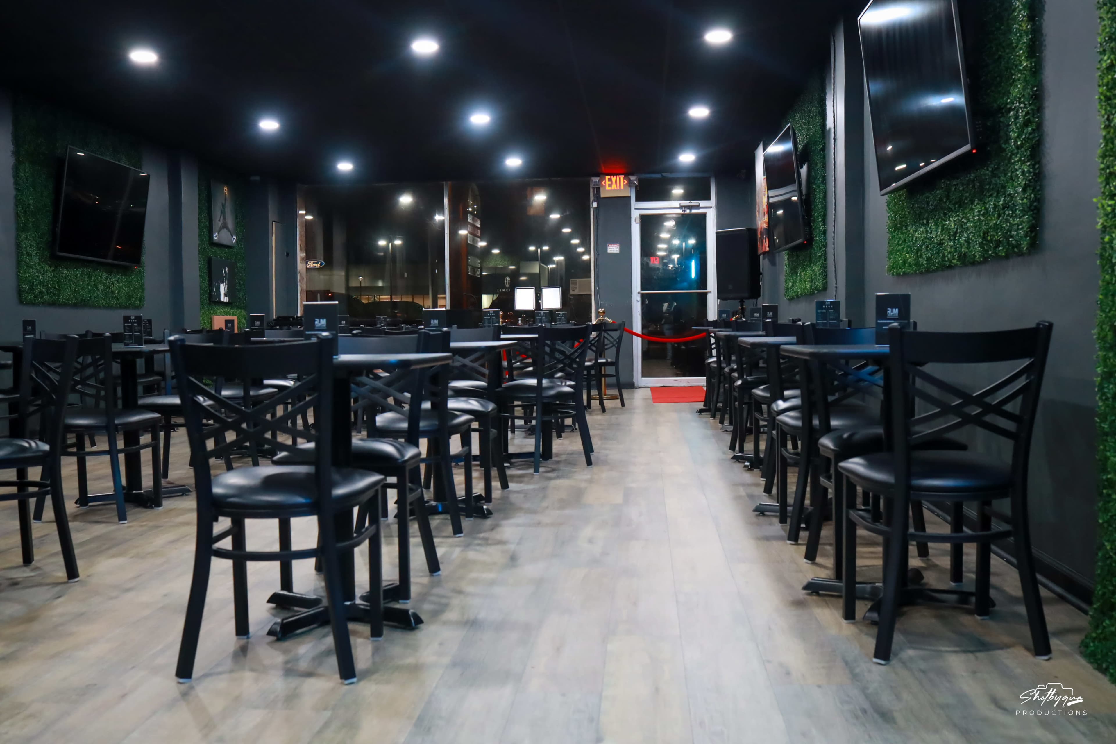 An indoor dining area with several black wooden tables and chairs arranged in rows on a wooden floor, surrounded by green wall installations and multiple television screens.
