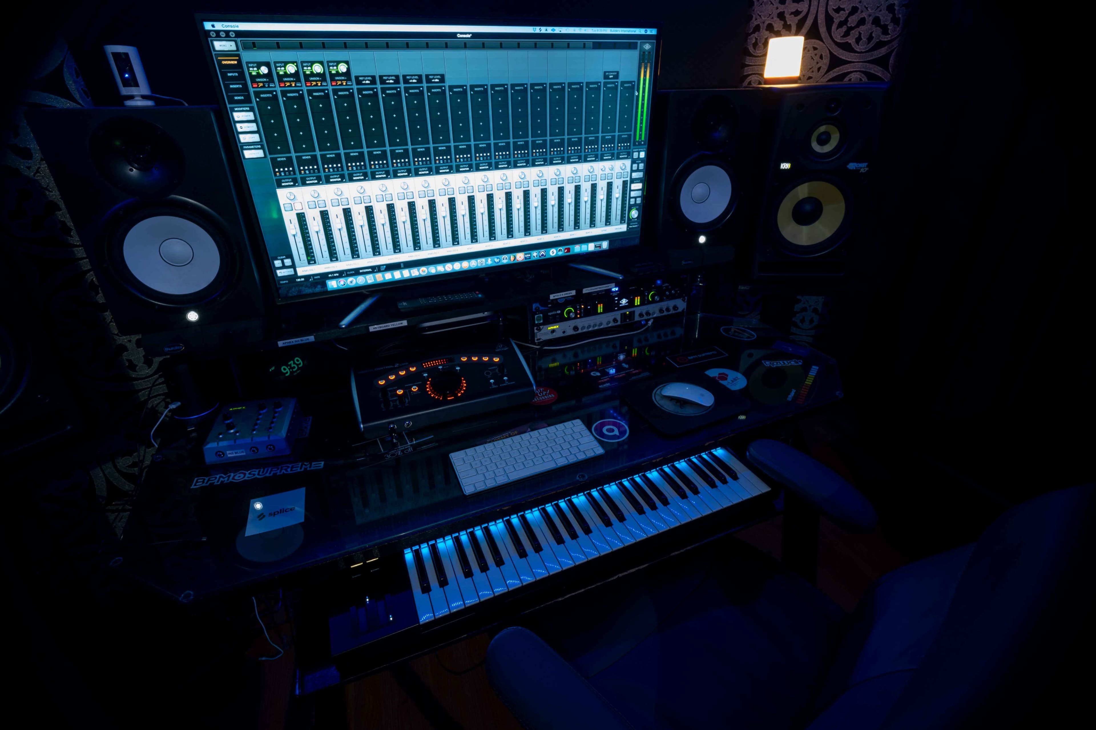 The image shows a dimly lit music studio setup featuring a large computer screen displaying a digital audio workstation, audio speakers, a MIDI keyboard, and various music production equipment on a desk.