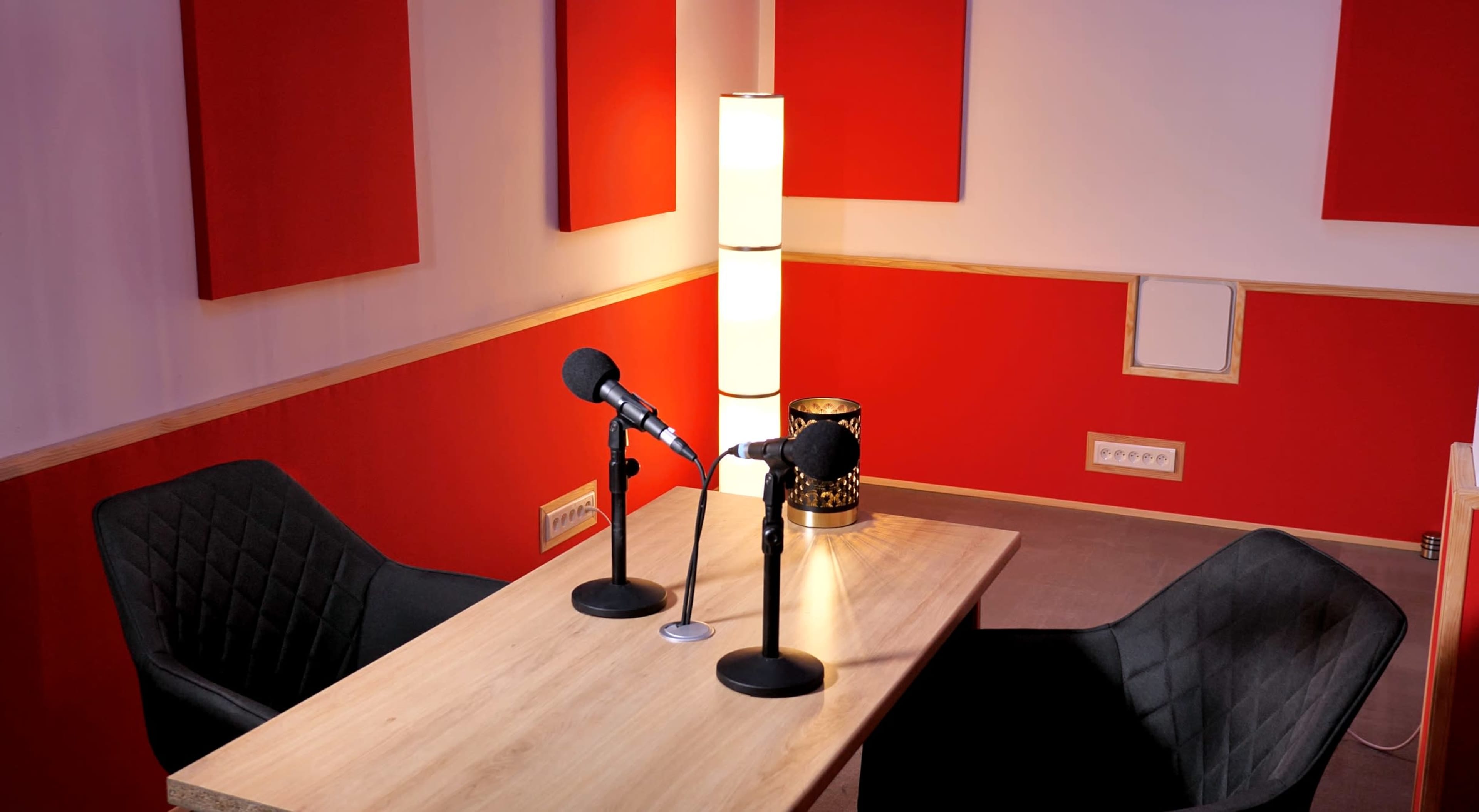 A podcast recording studio features a wooden table with two microphones, two chairs, and red acoustic panels on the walls.