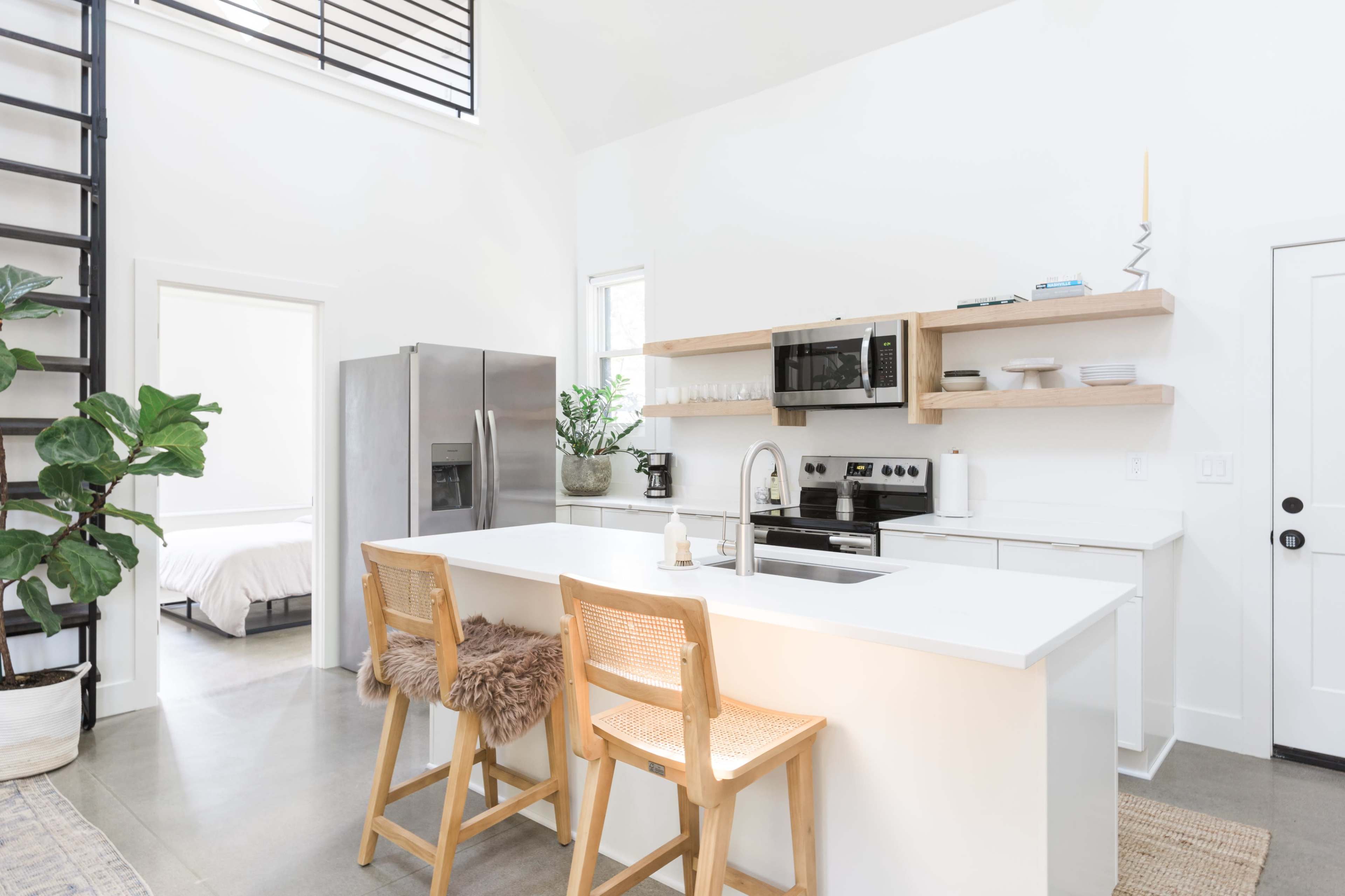 A modern kitchen features a white island with two wooden stools, stainless steel appliances, open shelving, and large windows allowing natural light.