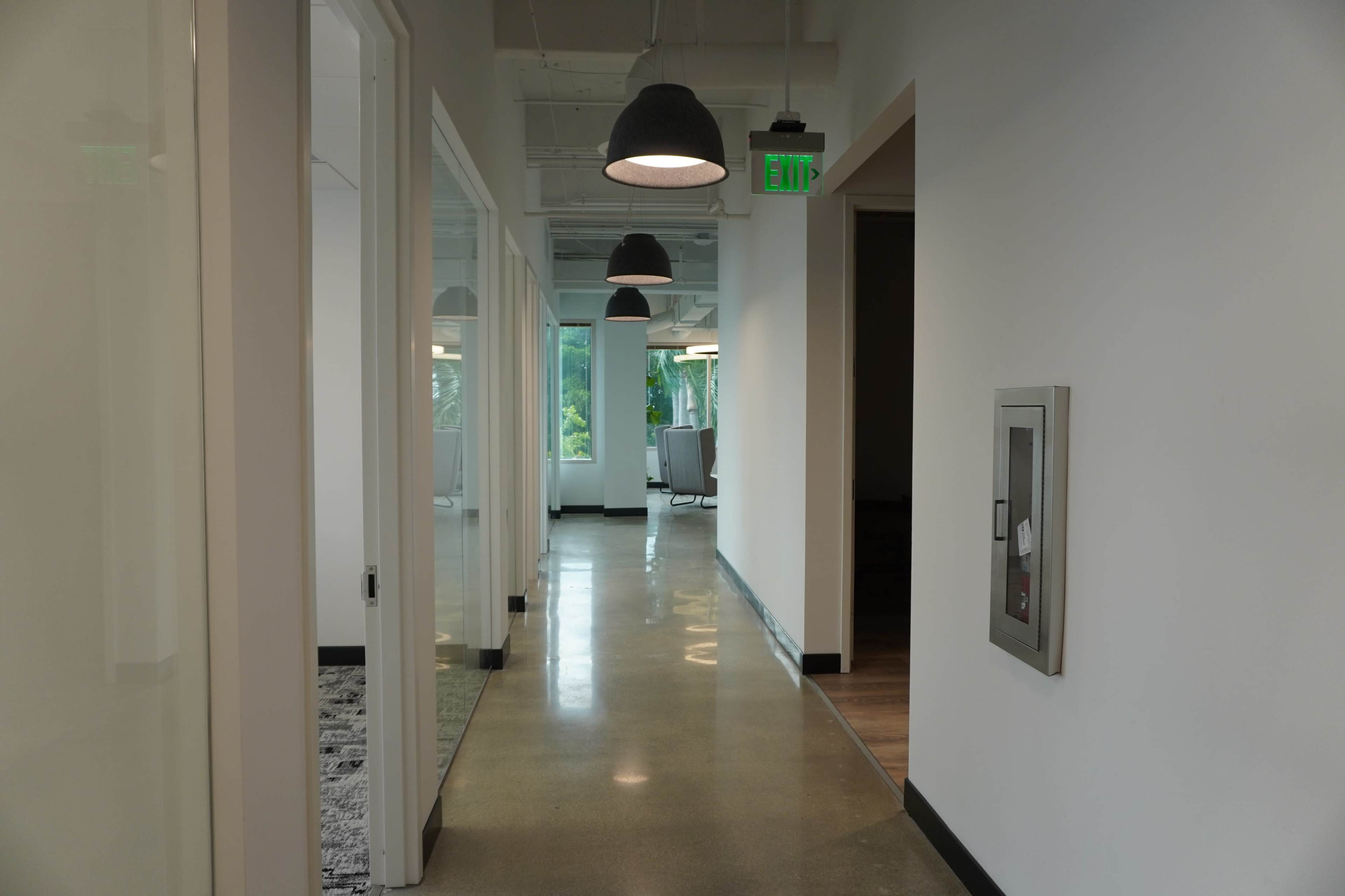 The image shows a modern office hallway with polished concrete floors, several pendant lights hanging from the ceiling, and glass-walled meeting rooms on either side.