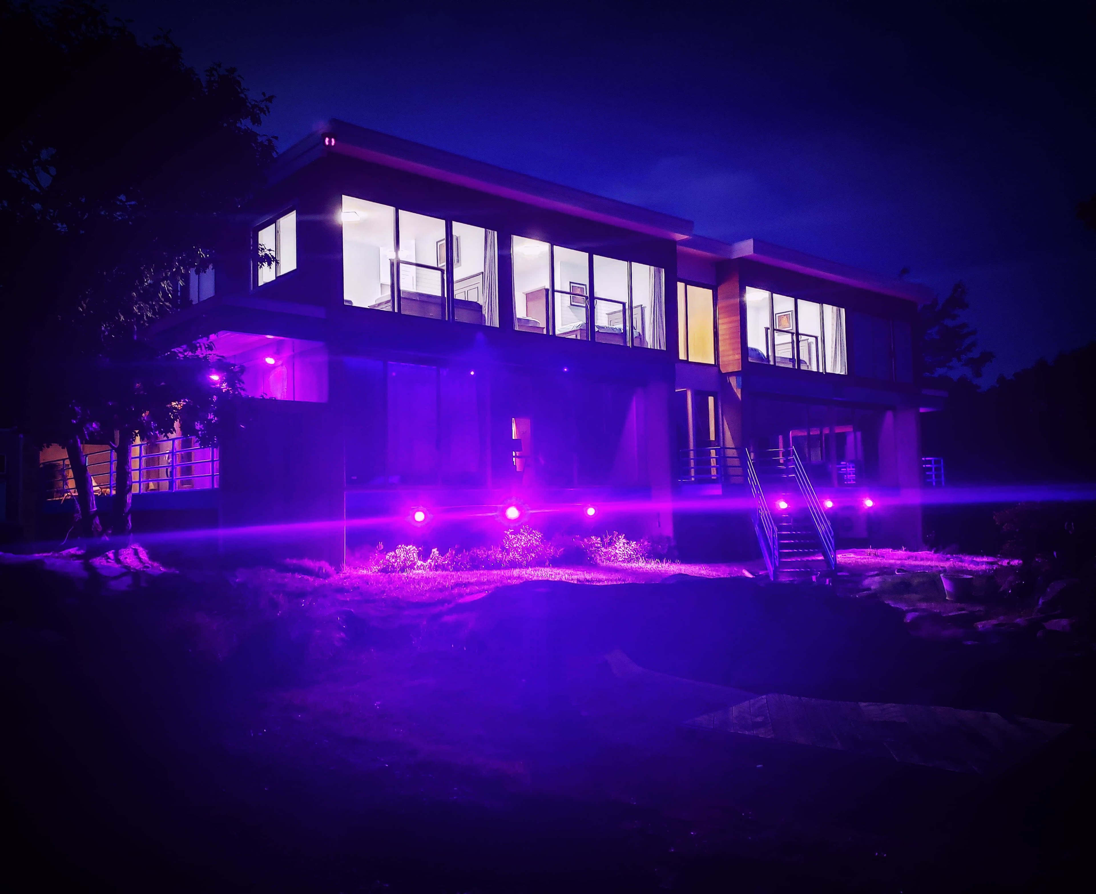 A modern house with large glass windows is illuminated by purple lights in a dark setting.