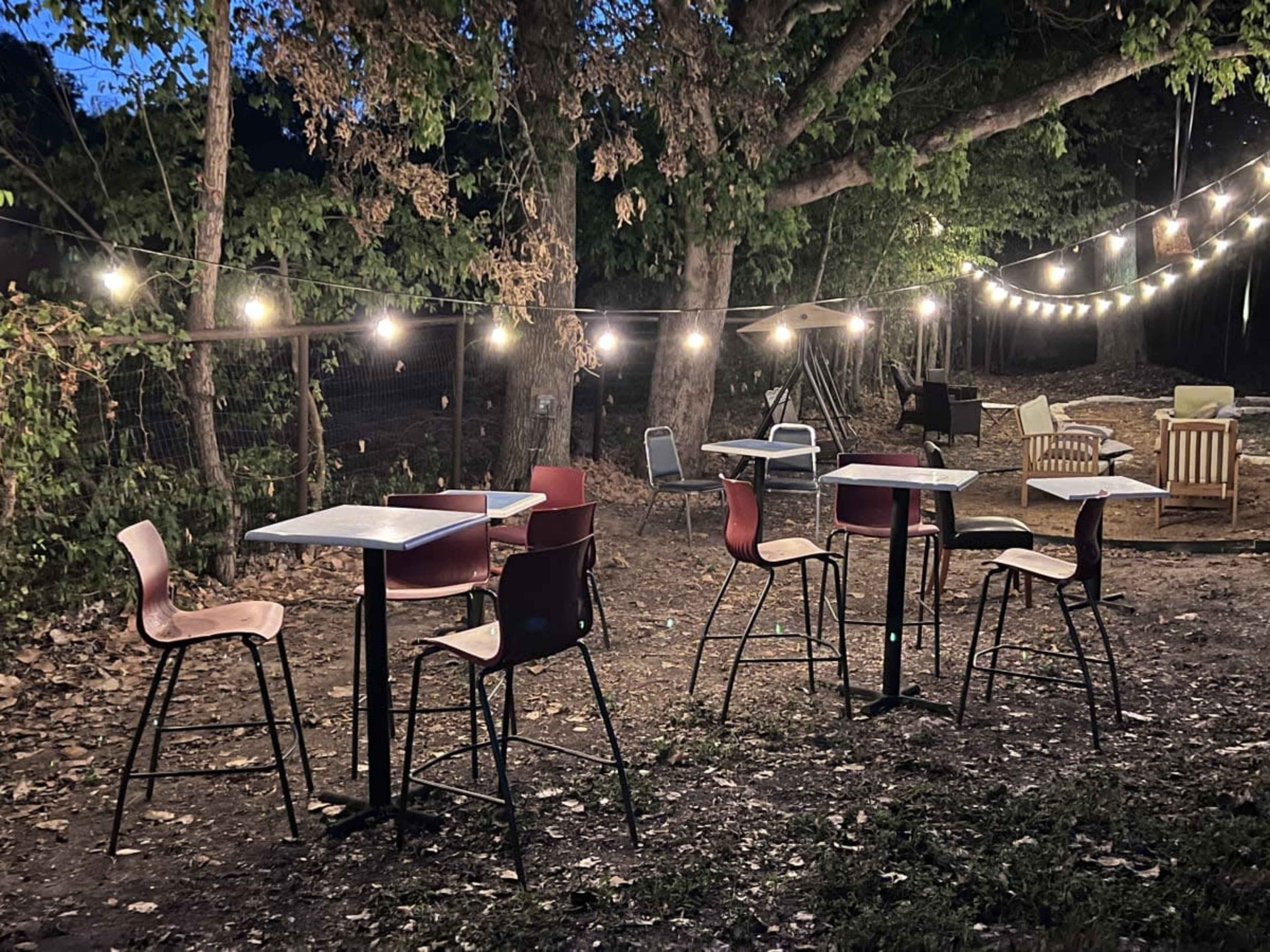 A small outdoor seating area with several tables and chairs is illuminated by string lights in a wooded setting.