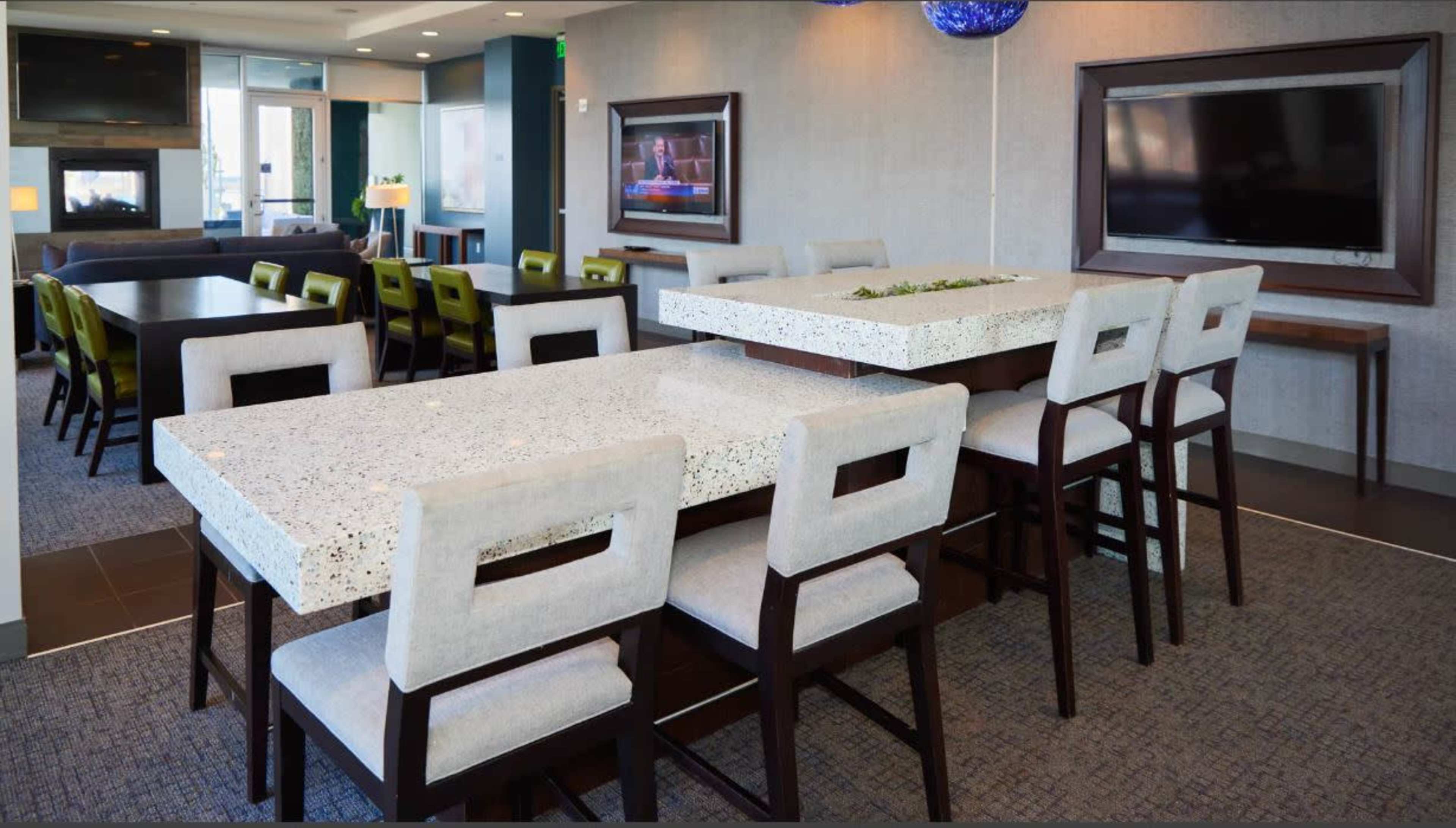 A modern dining area with a large white terrazzo table surrounded by upholstered chairs, with additional seating and large windows in the background.