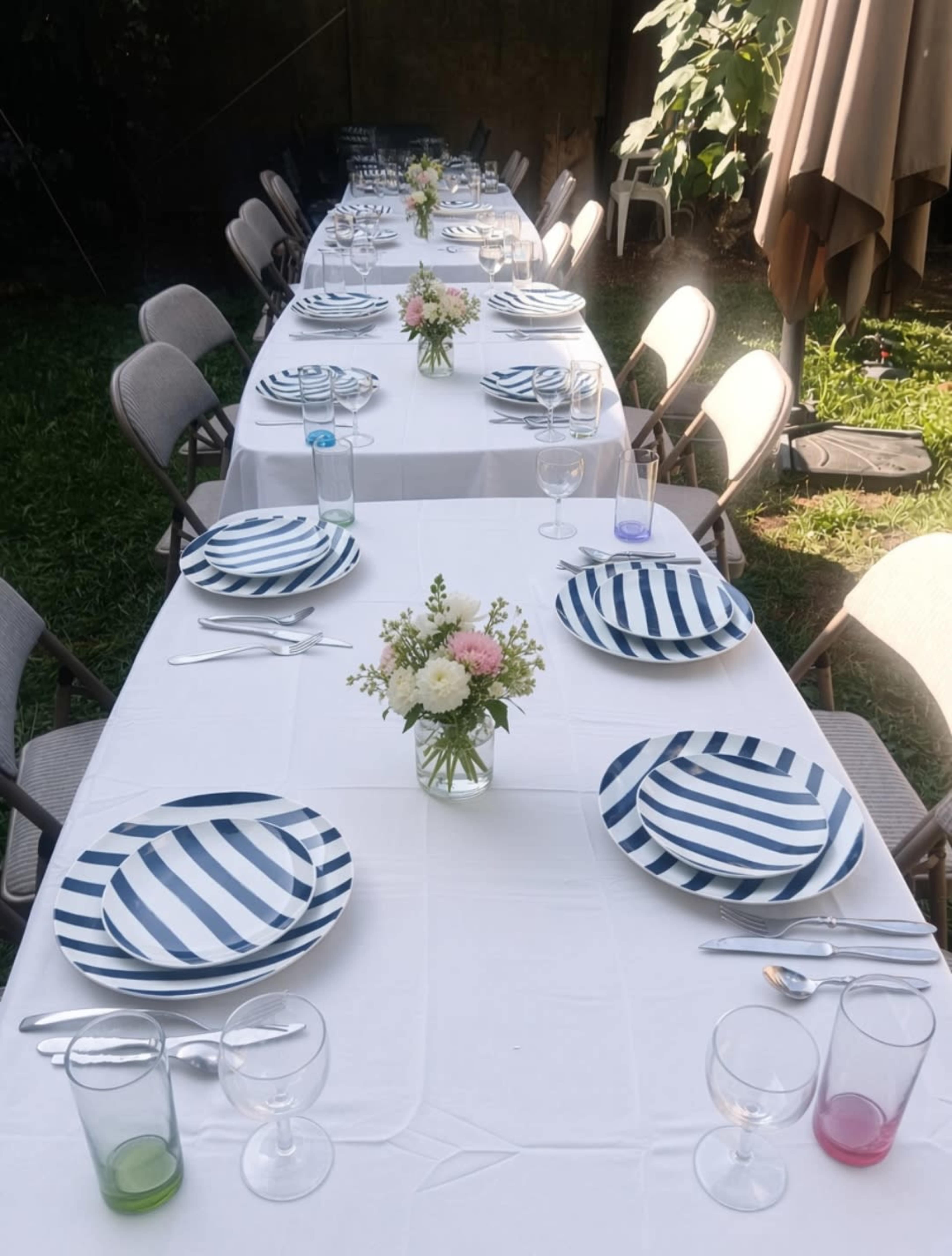 A long table is set outdoors with striped plates, glasses, and small floral arrangements, surrounded by folding chairs.