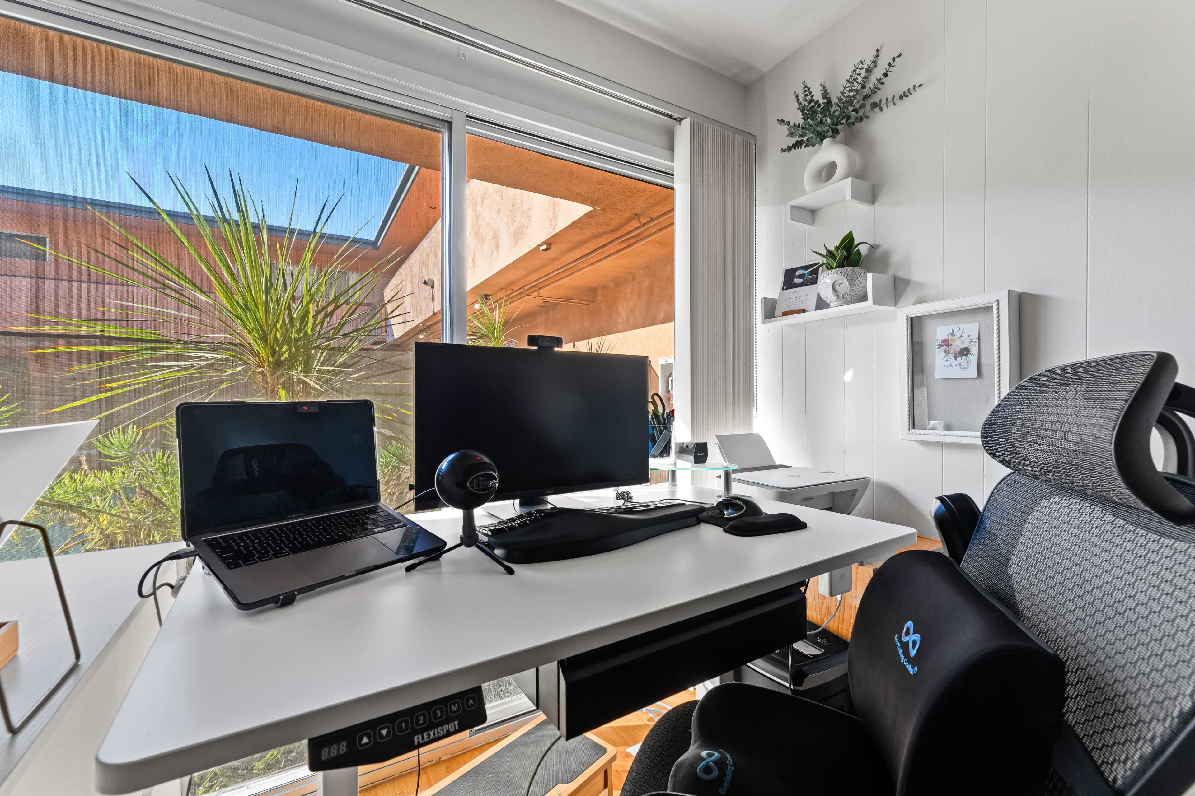 A modern home office setup features a desk with a laptop, monitor, and headset, positioned next to a large window that frames a view of palm trees.