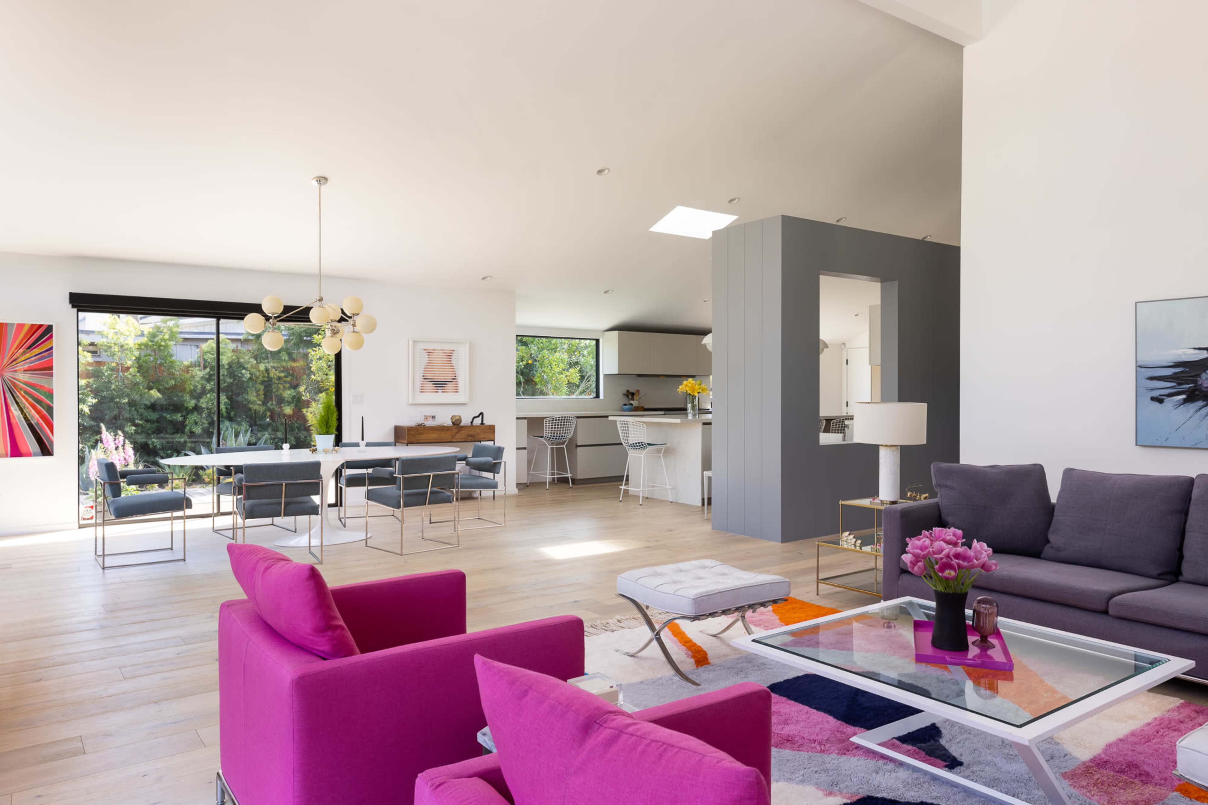 A modern living room with a pink sofa, glass coffee table, and an open layout connecting to a dining area and kitchen with large windows.