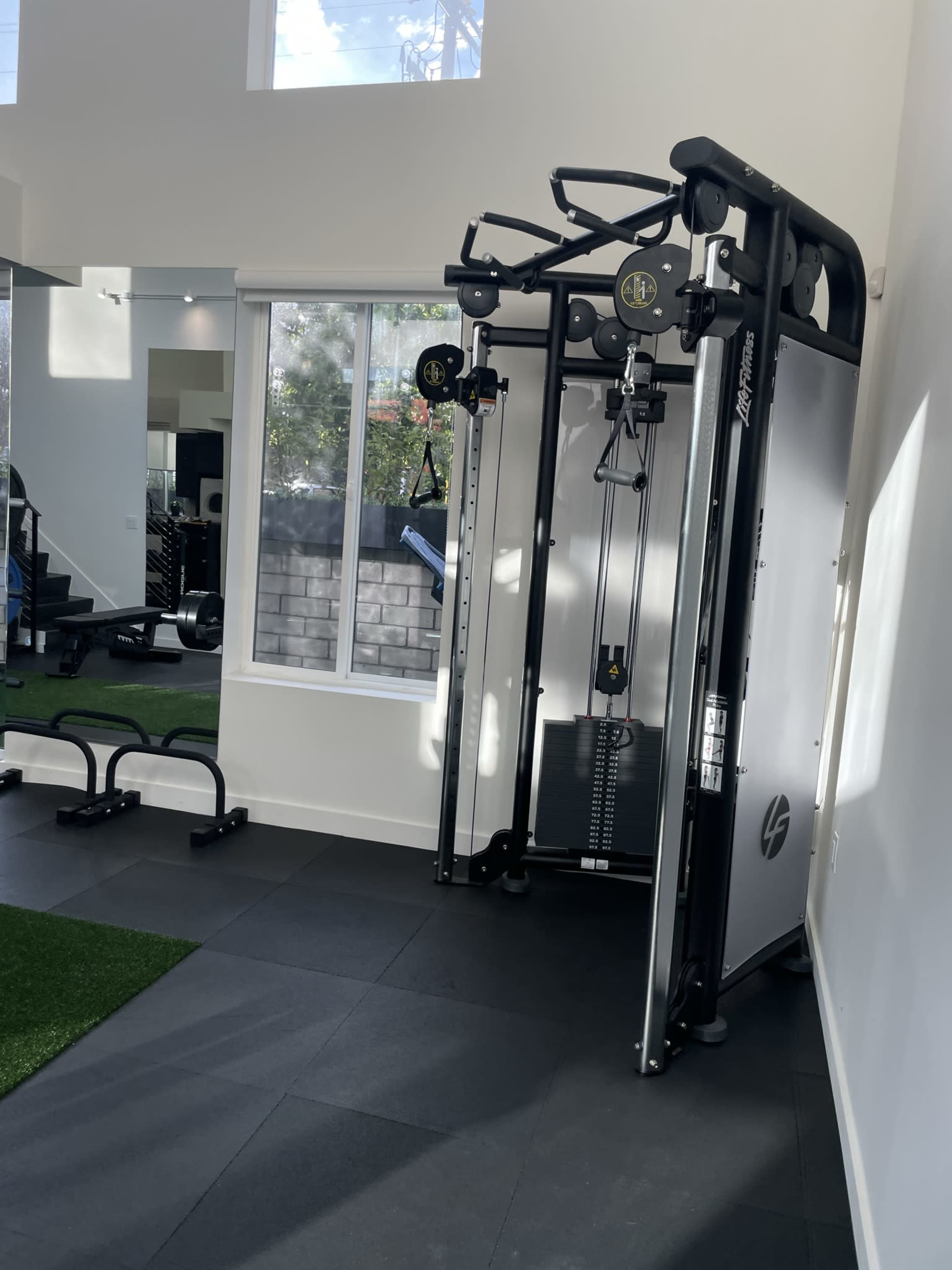 A home gym equipped with a cable machine in a well-lit room, featuring large windows and a rubber flooring area.