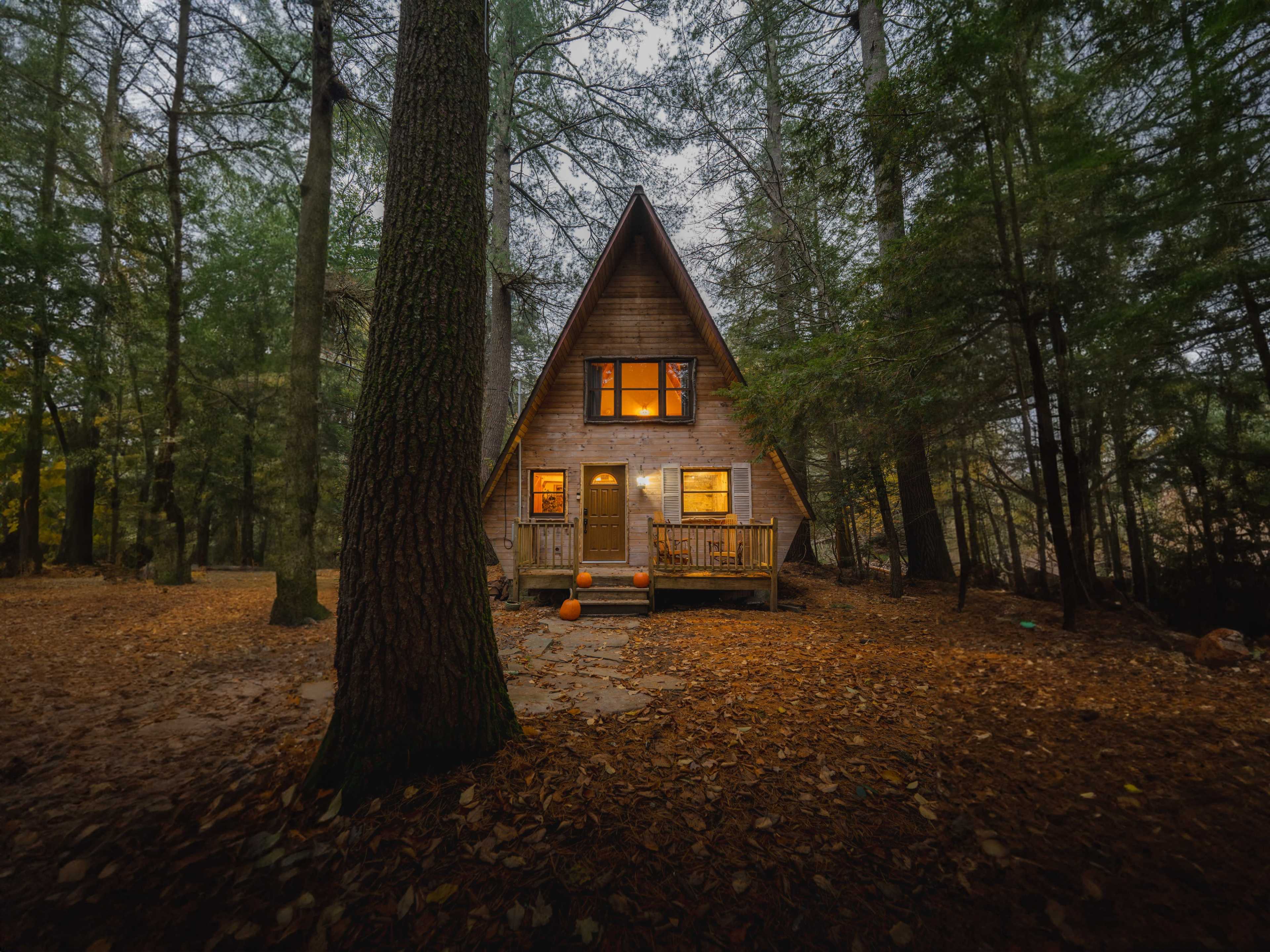 An A-frame cabin with illuminated windows sits among trees covered in fallen leaves.