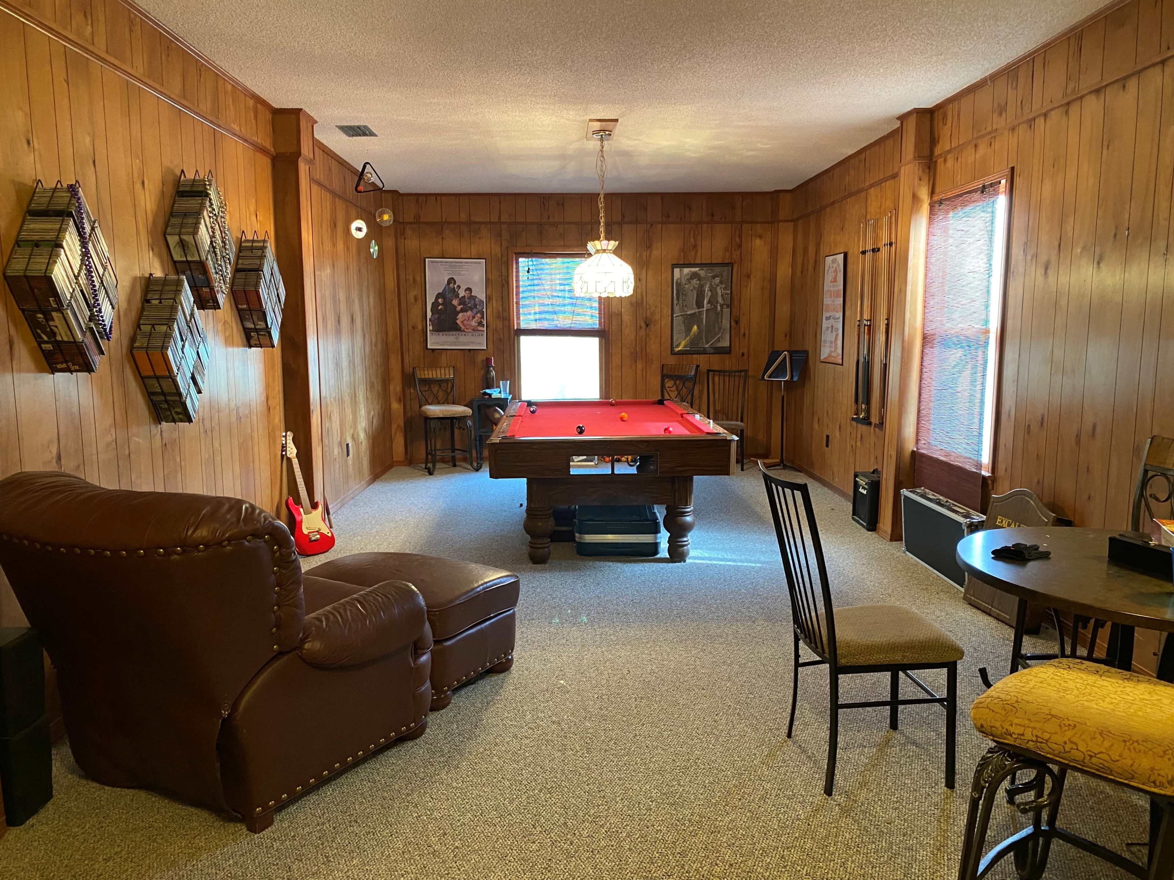 The room features wooden paneling, a pool table at the center, and various seating arrangements, including a large recliner and chairs.