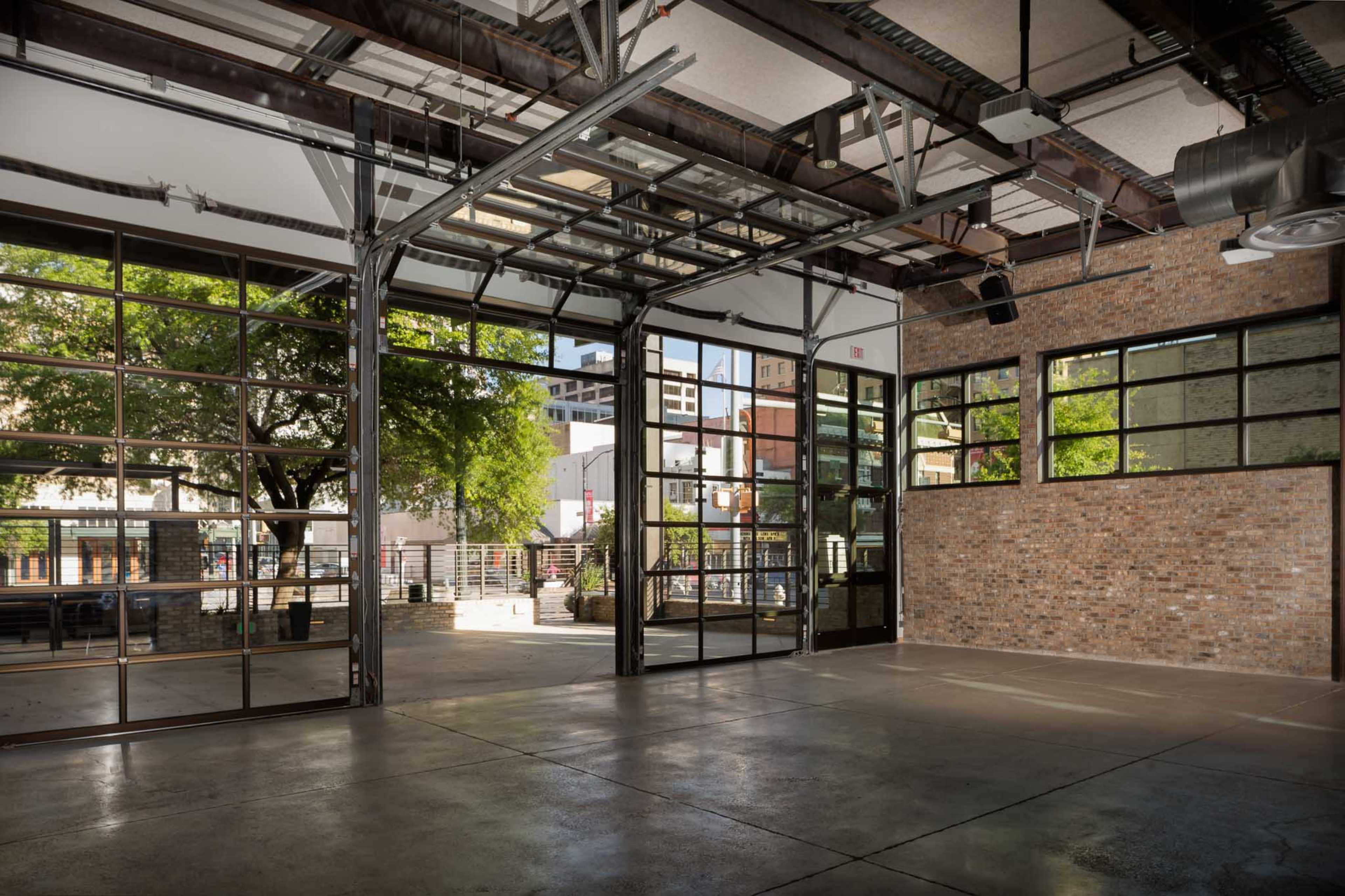 An industrial-style interior space with large glass doors and a brick wall, featuring an open layout and trees visible outside.