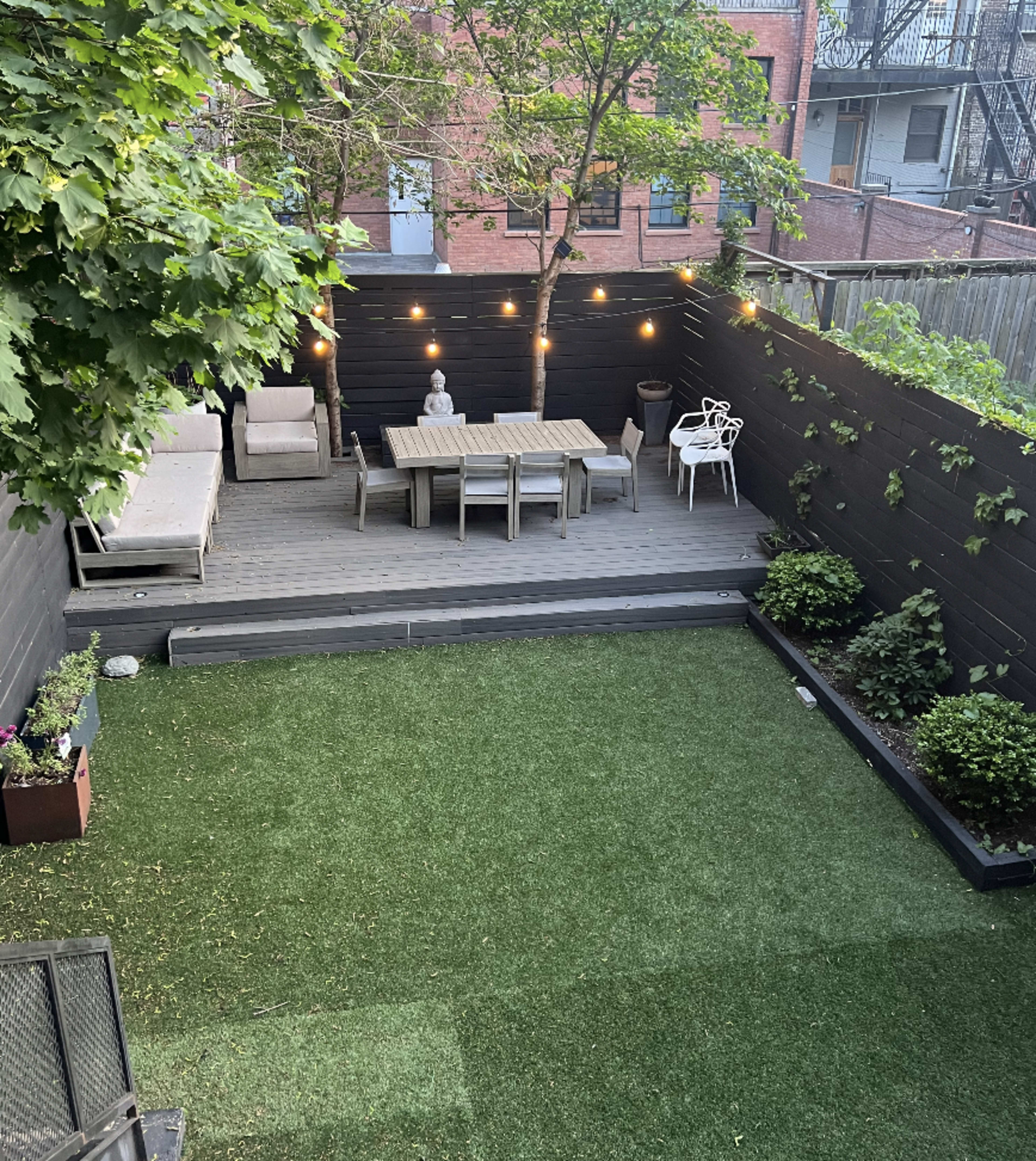 A landscaped backyard features a dining table surrounded by chairs, decorative plants, a stone statue, and string lights overhead.