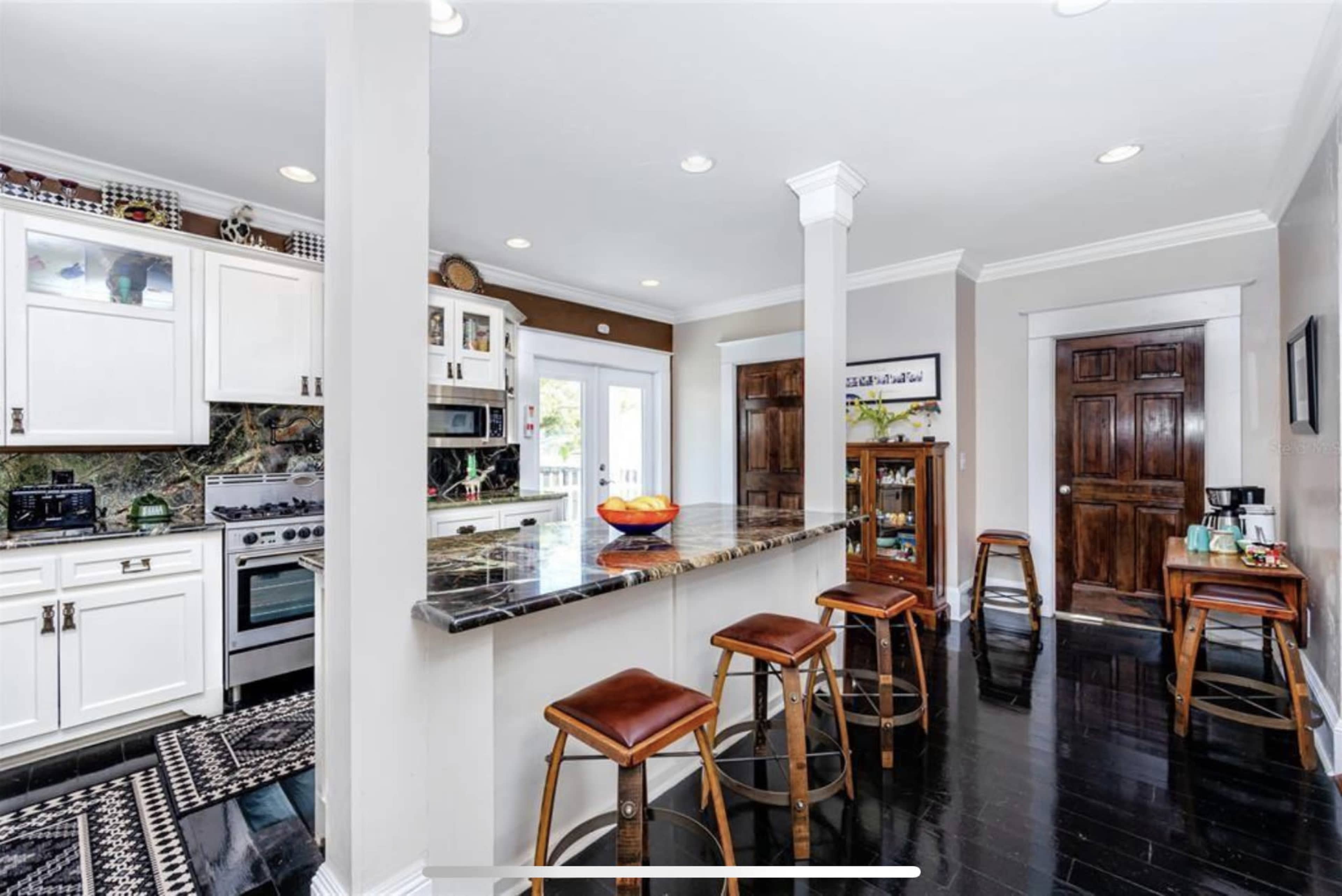 The image shows a modern kitchen with white cabinetry, a granite countertop, and bar stools, with a view of an outdoor area through a window.