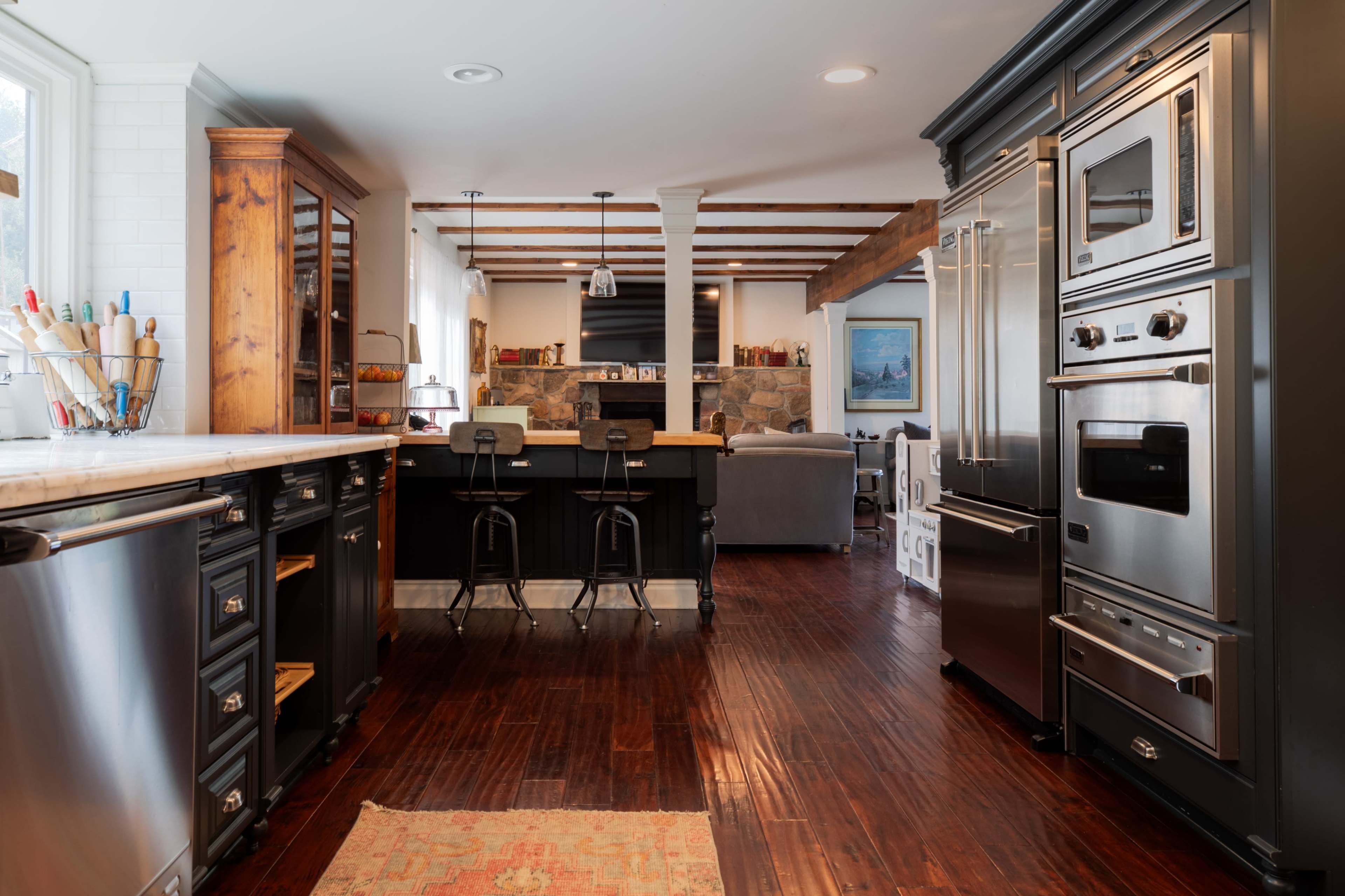 The image shows a modern kitchen featuring stainless steel appliances, dark cabinetry, and a wooden dining area with bar stools.