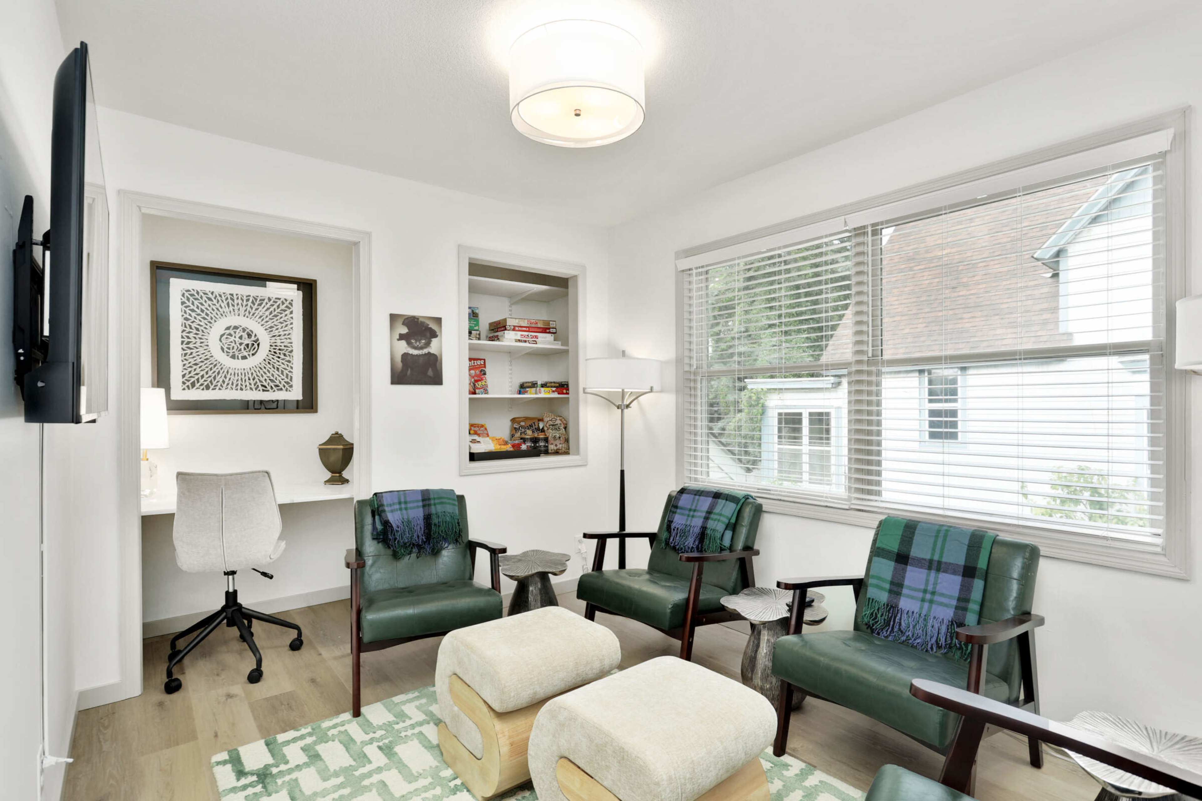 A neatly arranged sitting area with two green chairs, a desk with a white chair, and a light fixture overhead, accompanied by a decorative lamp and ottomans.