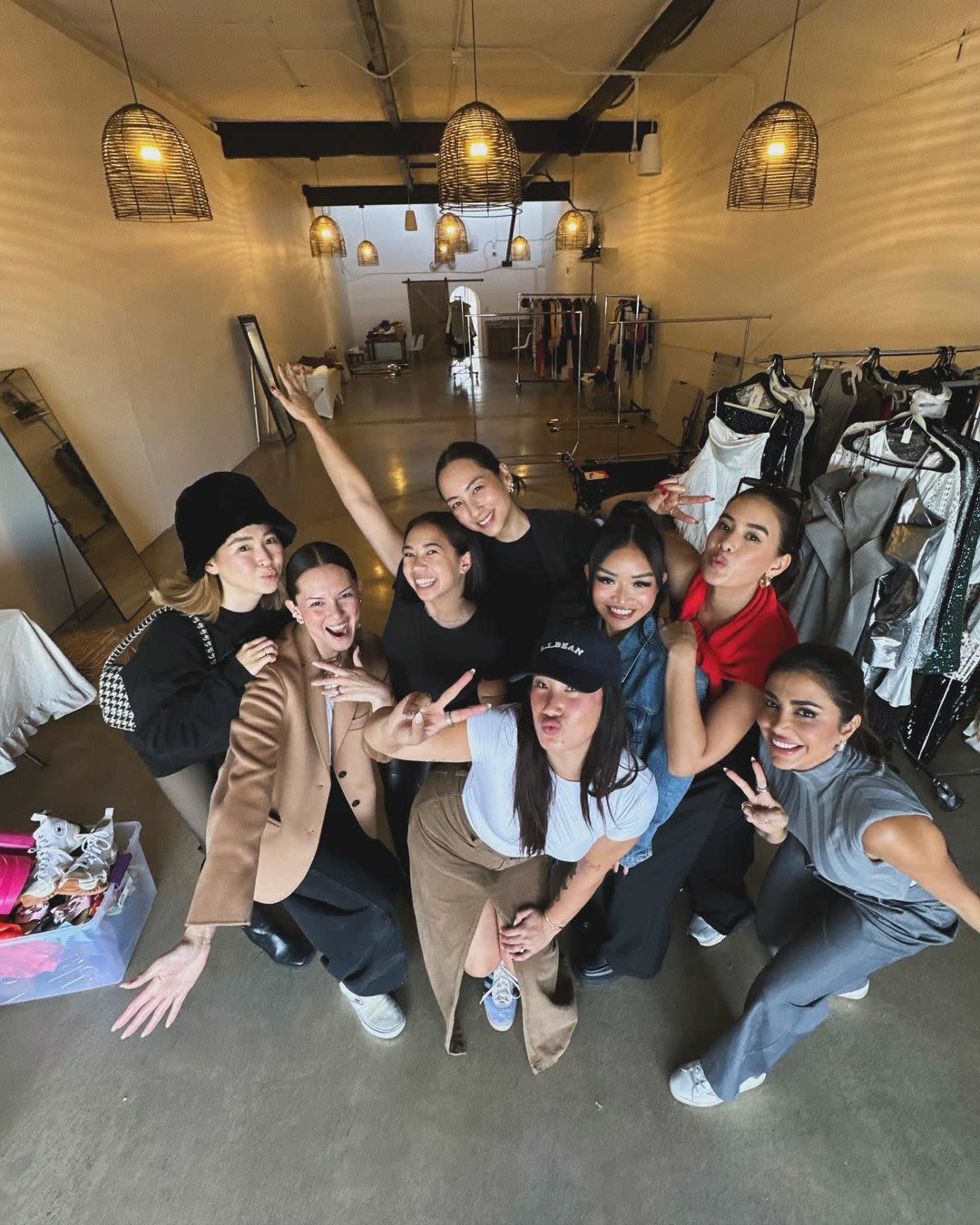 A group of eight women poses playfully in a spacious, well-lit studio filled with clothing racks and mirrors.