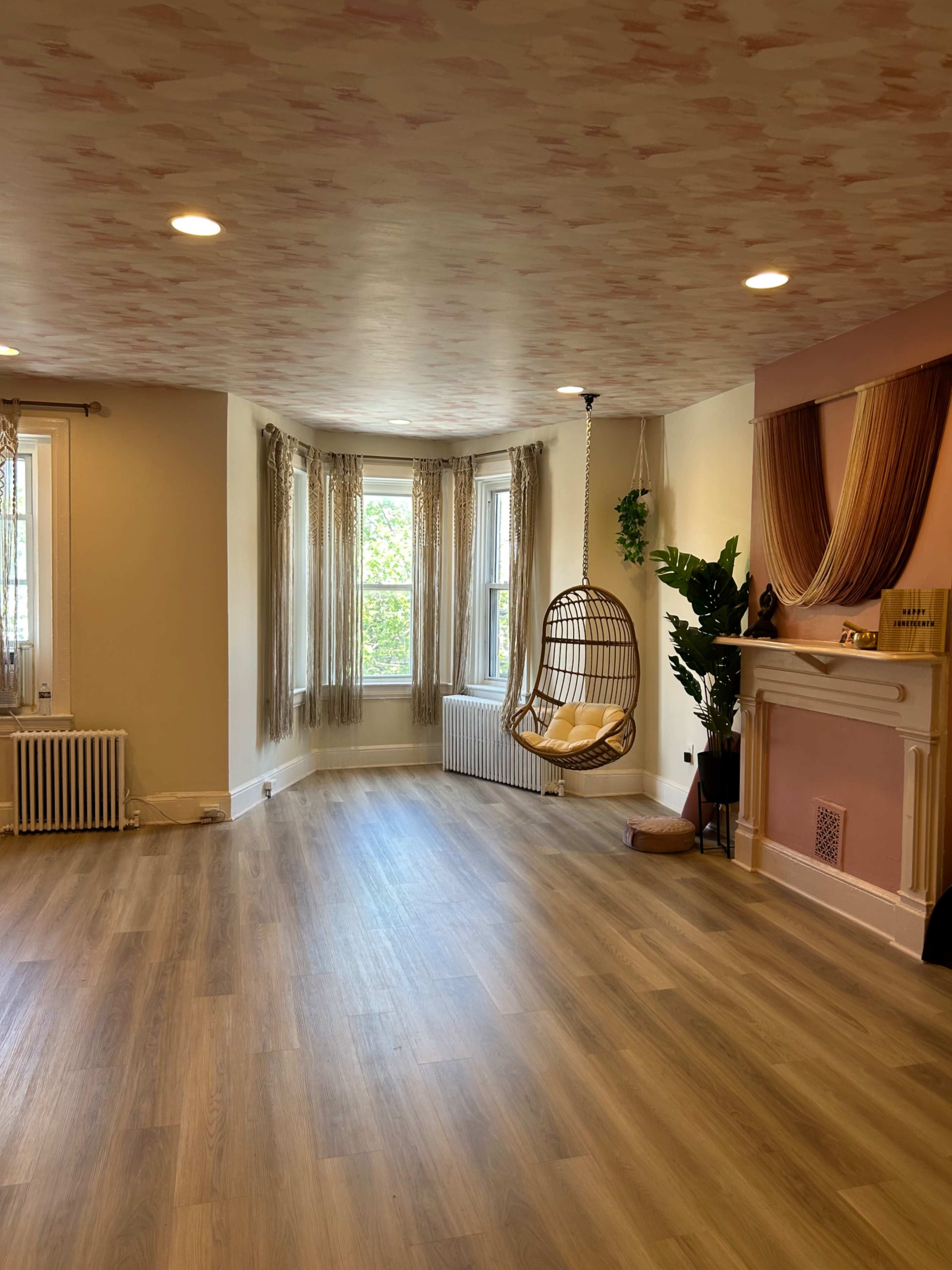The image shows a spacious room with a hanging wicker chair, large windows, and a fireplace adorned with decorative fabric.