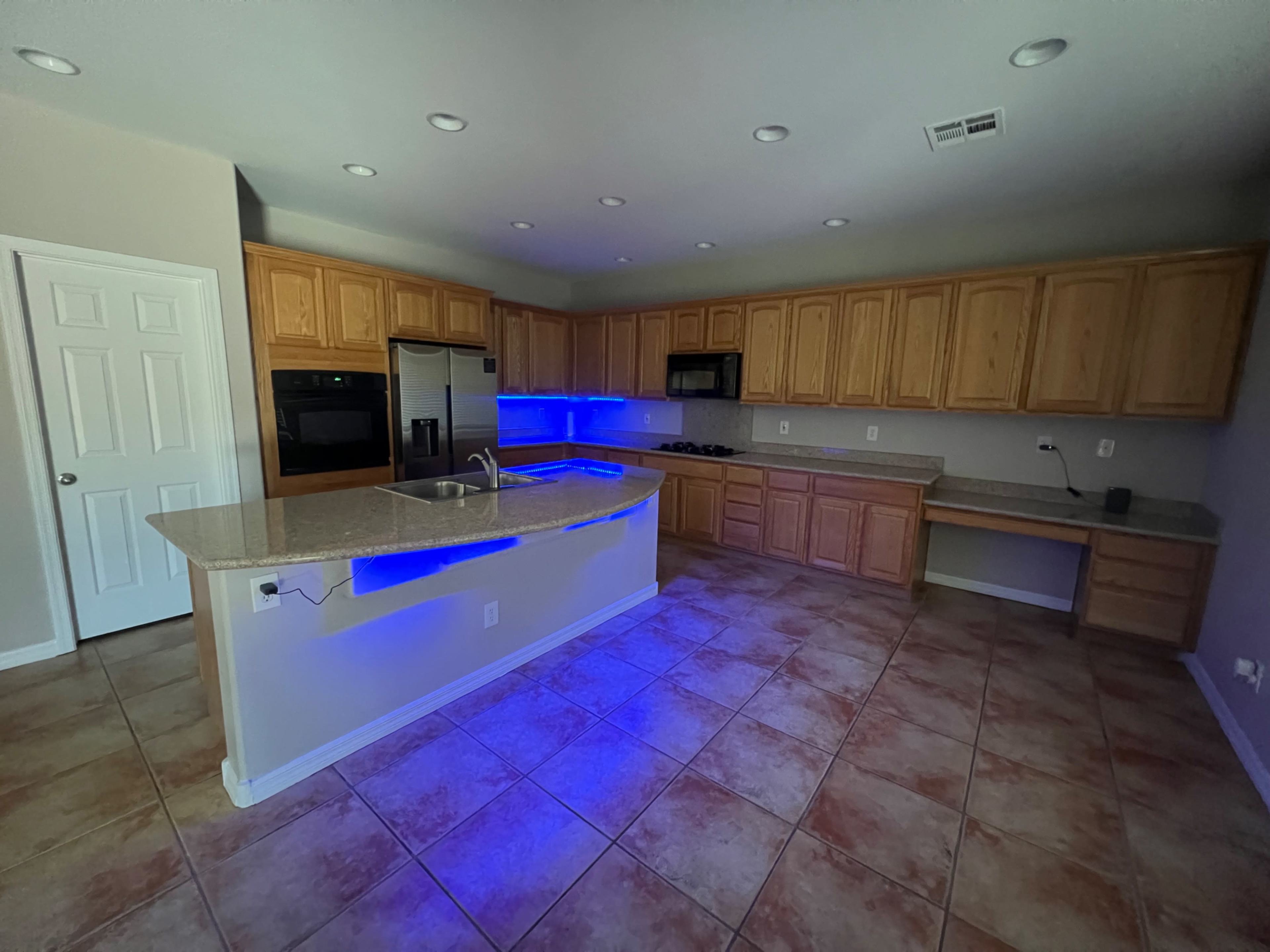 A kitchen features wooden cabinets, a granite countertop with an undermount sink, and blue lighting along the bar area.