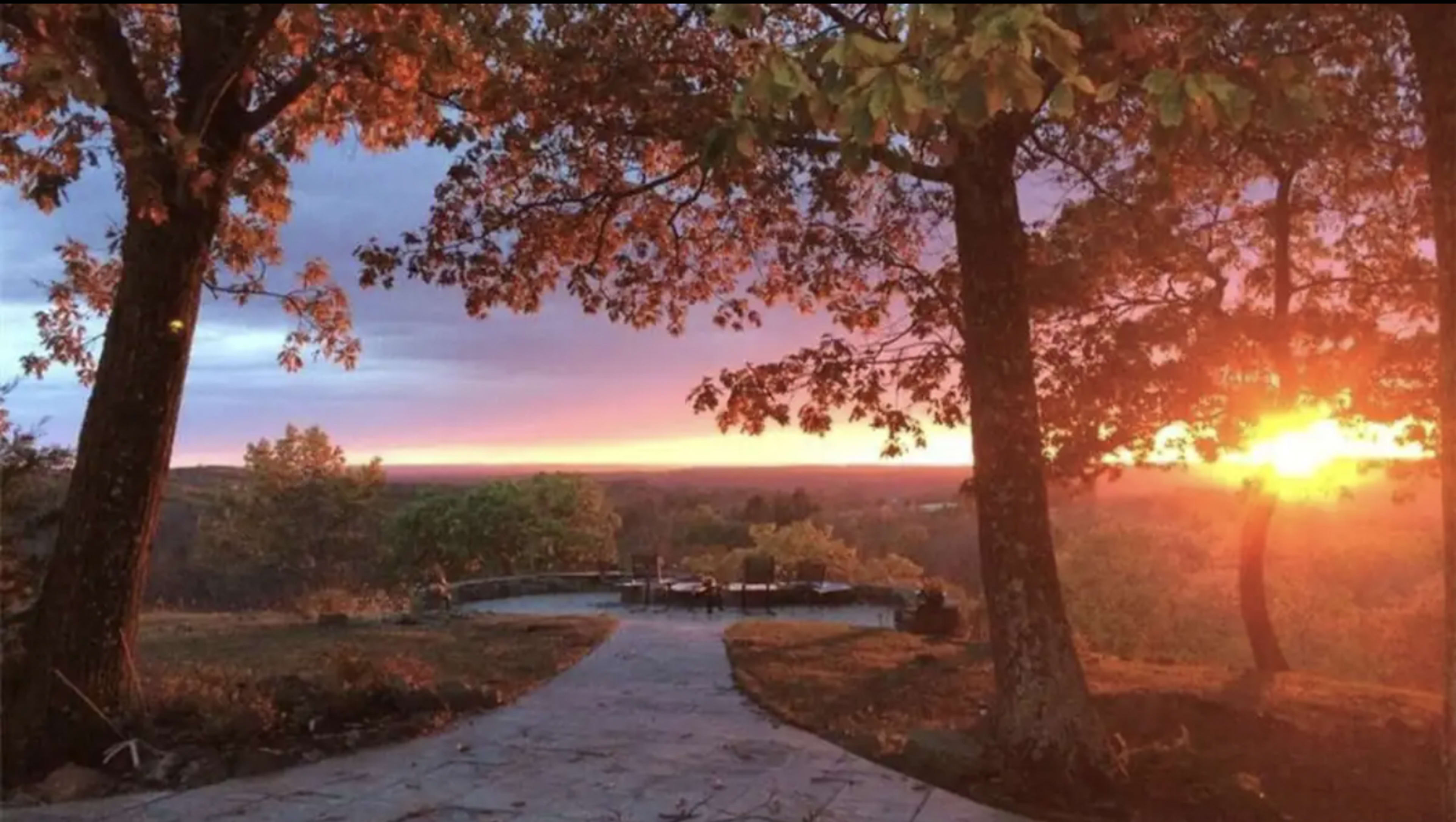 The scene shows a pathway leading through trees to a sunset over a valley, with a patio area visible in the distance.