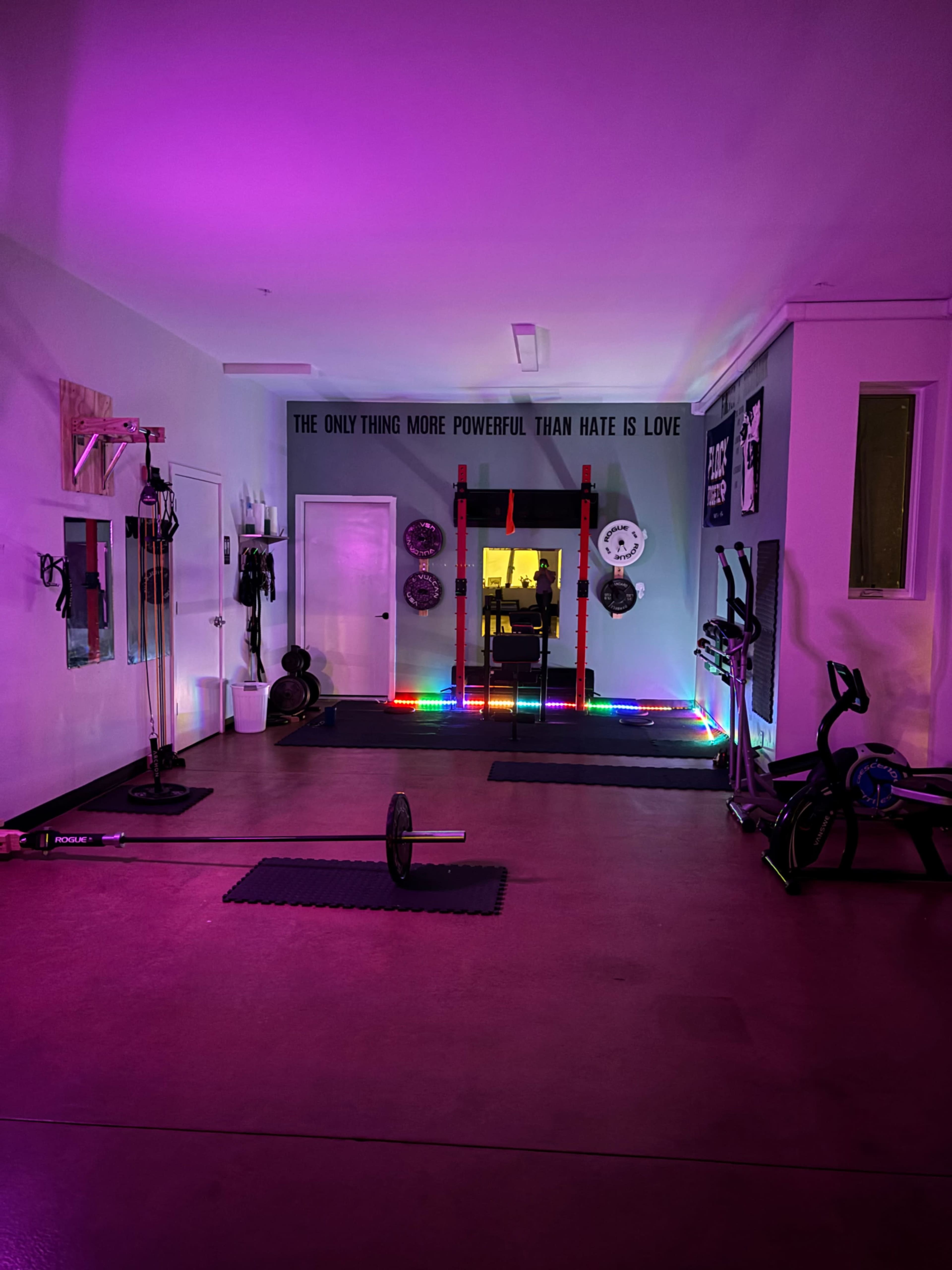 A brightly lit home gym featuring colorful LED lights, a barbell on the floor, and various fitness equipment arranged around a mirrored wall.