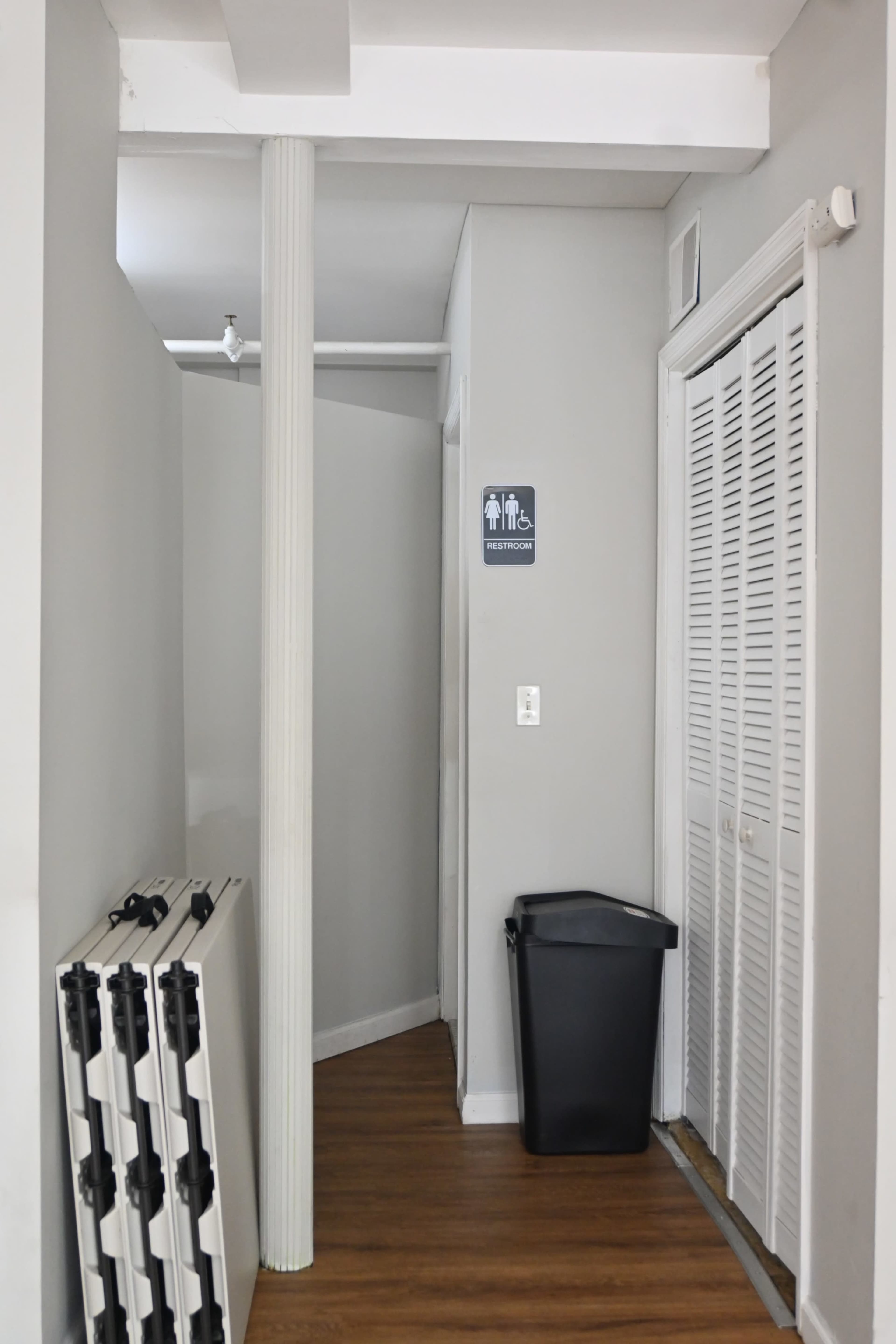 The image shows a narrow hallway leading to a door with an accessible restroom sign, flanked by a white column and a trash can, with folding tables stacked against the wall.