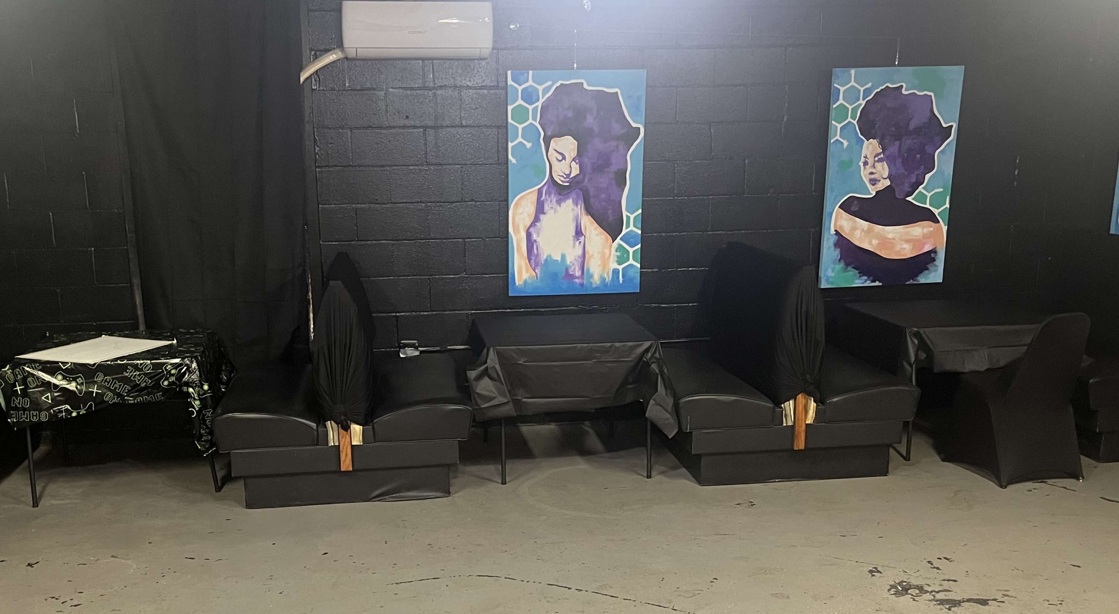 The scene shows a dimly lit room with black walls, featuring several black chairs and tables adorned with art pieces of women on the walls.