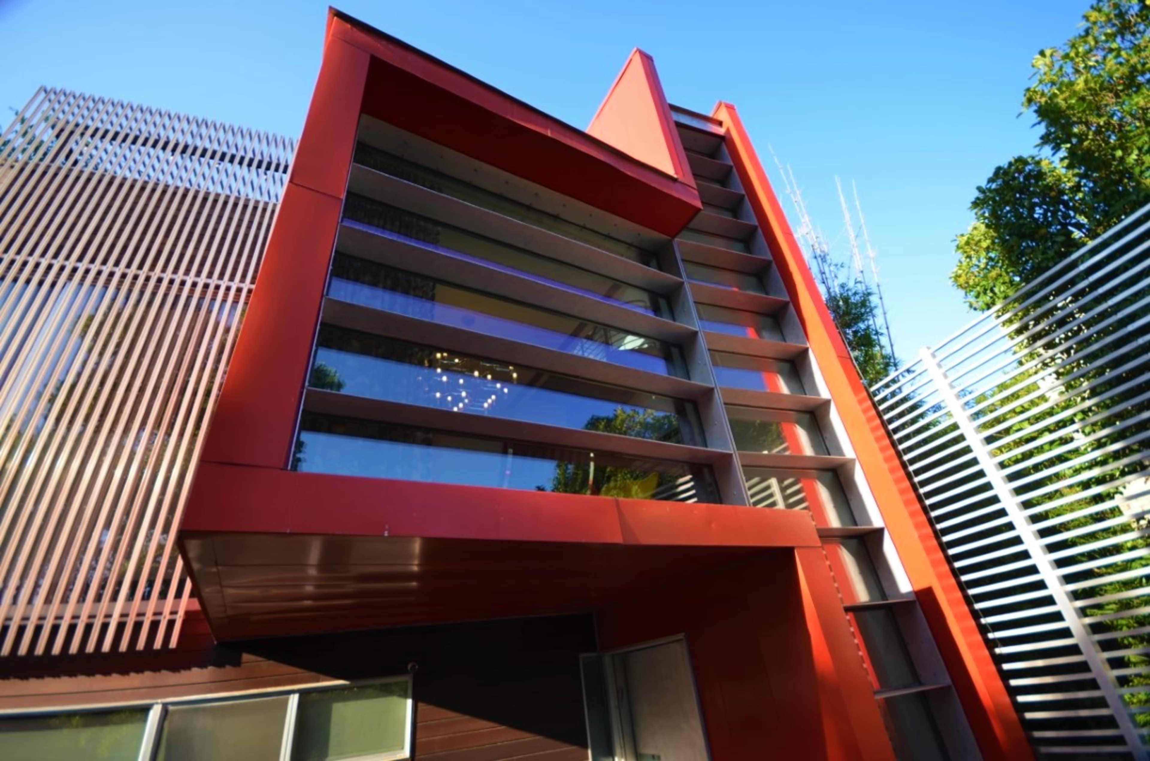 A modern red house with large windows and geometric lines, surrounded by trees and a metal fence.