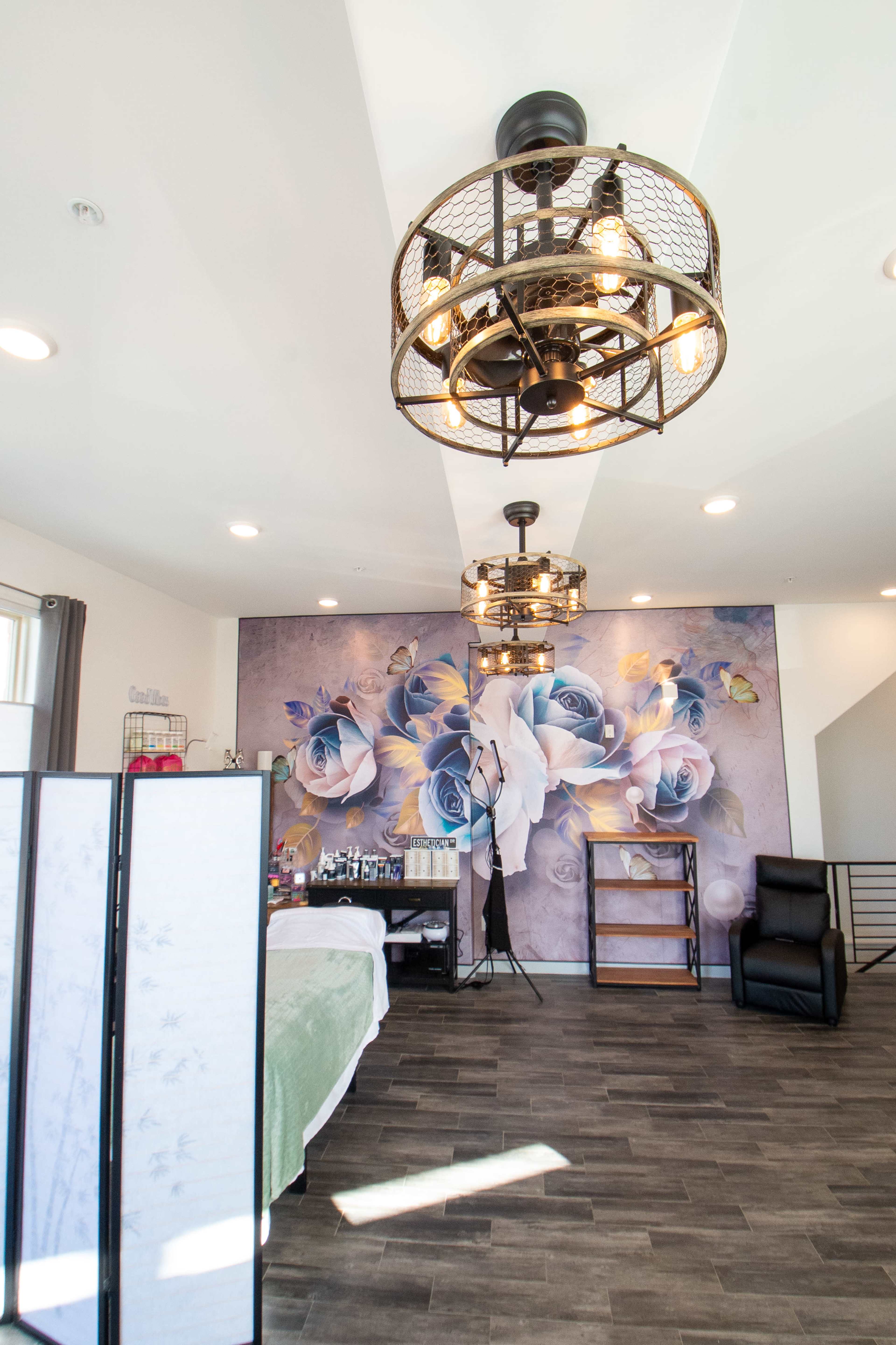 A modern spa interior featuring two industrial-style light fixtures, a floral mural on the wall, and a treatment bed with a partition.