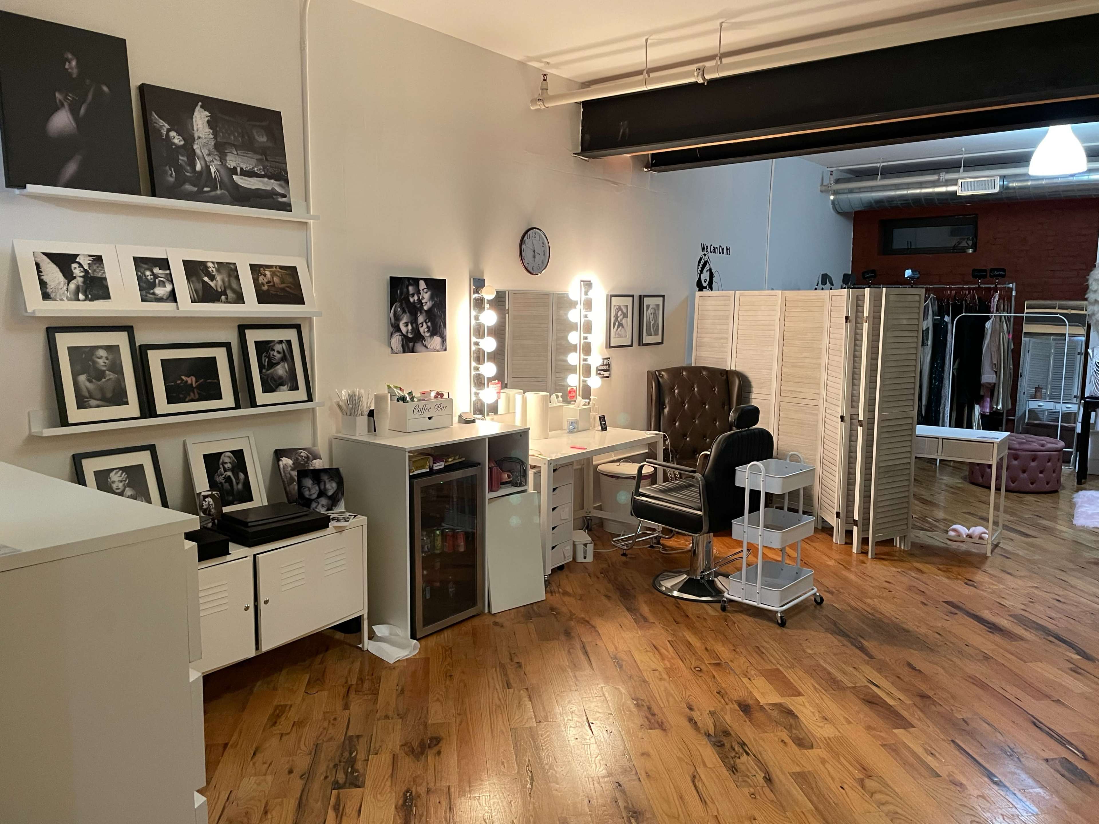 The image shows a spacious photography studio with framed black-and-white portraits on the walls, a well-lit makeup station, a large brown armchair, and a partition separating different areas.