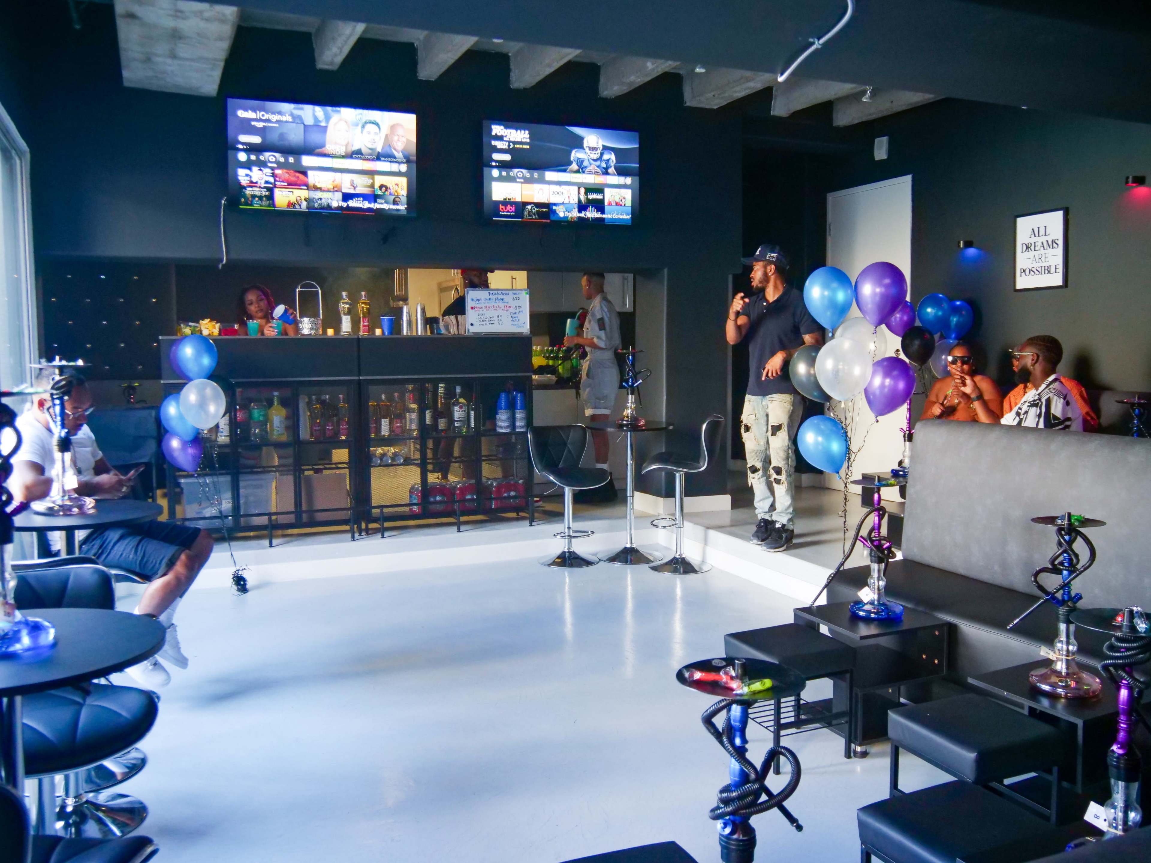 A modern lounge features multiple televisions, hookah setups, and patrons seated at tables, surrounded by balloons and a snack bar.