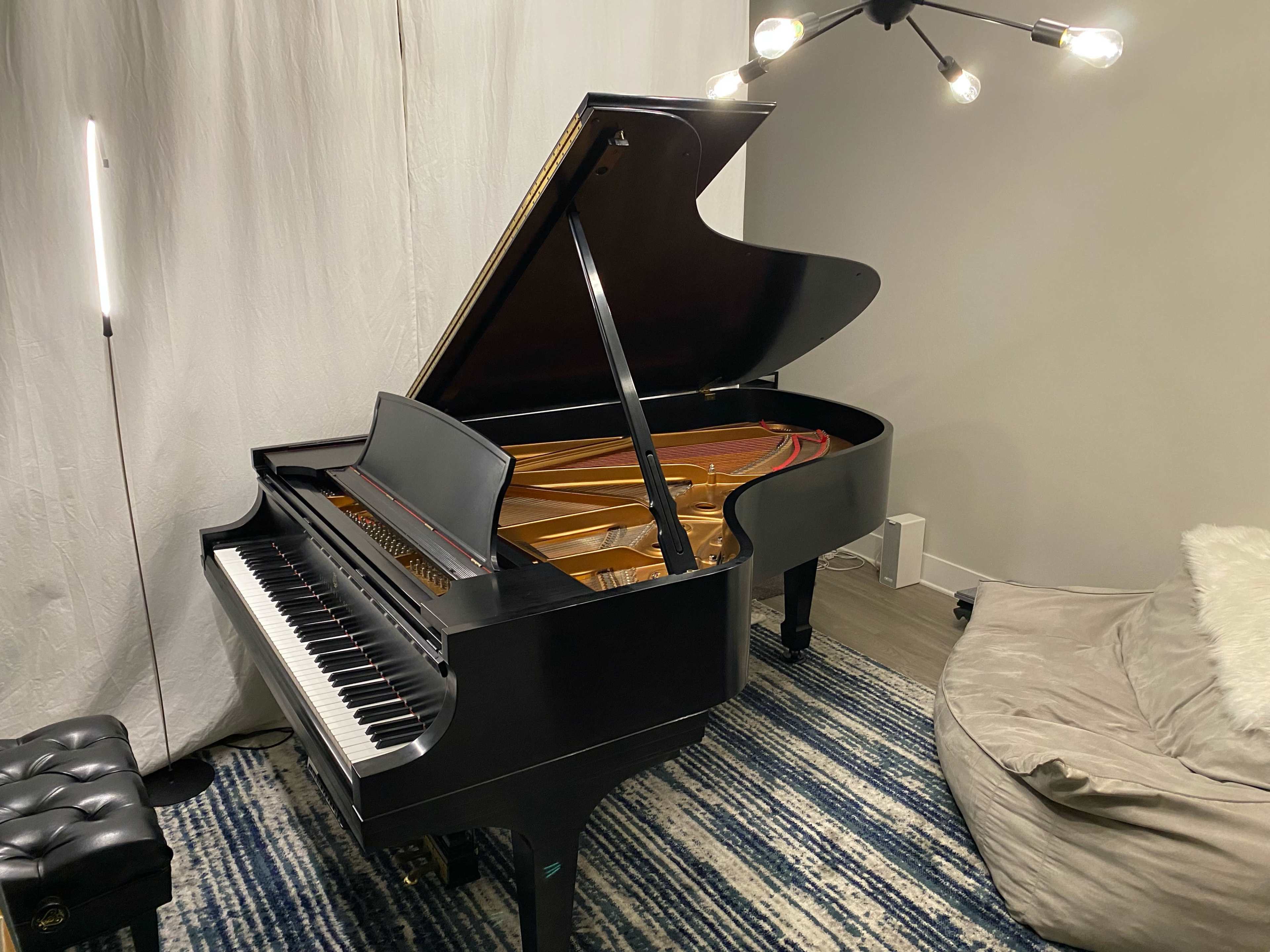 A grand piano with its lid open is positioned beside a soft couch and illuminated by a modern light fixture in a softly lit room.