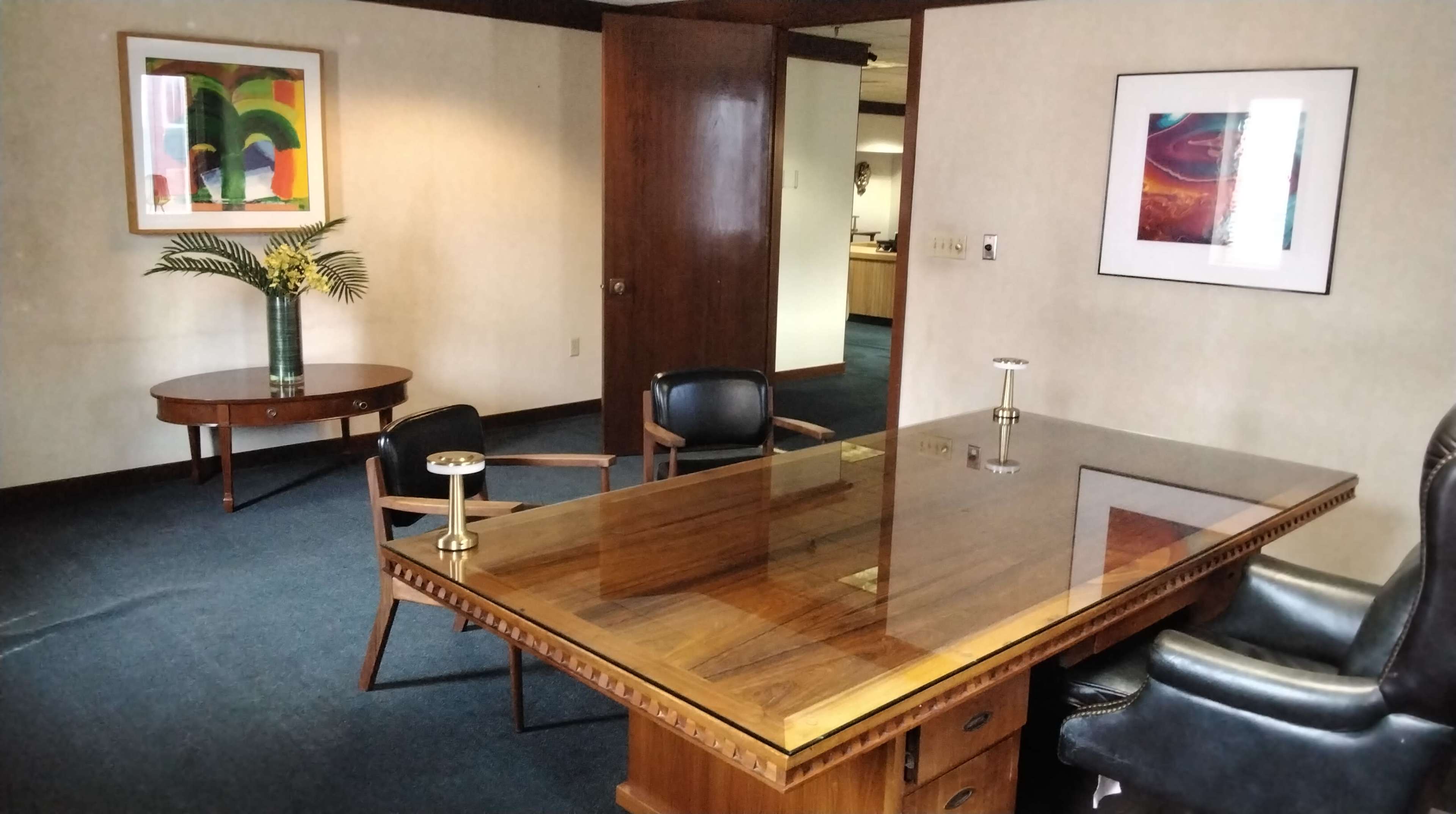 The image shows an office space featuring a wooden desk, two chairs, a floral arrangement, and artwork on the walls.