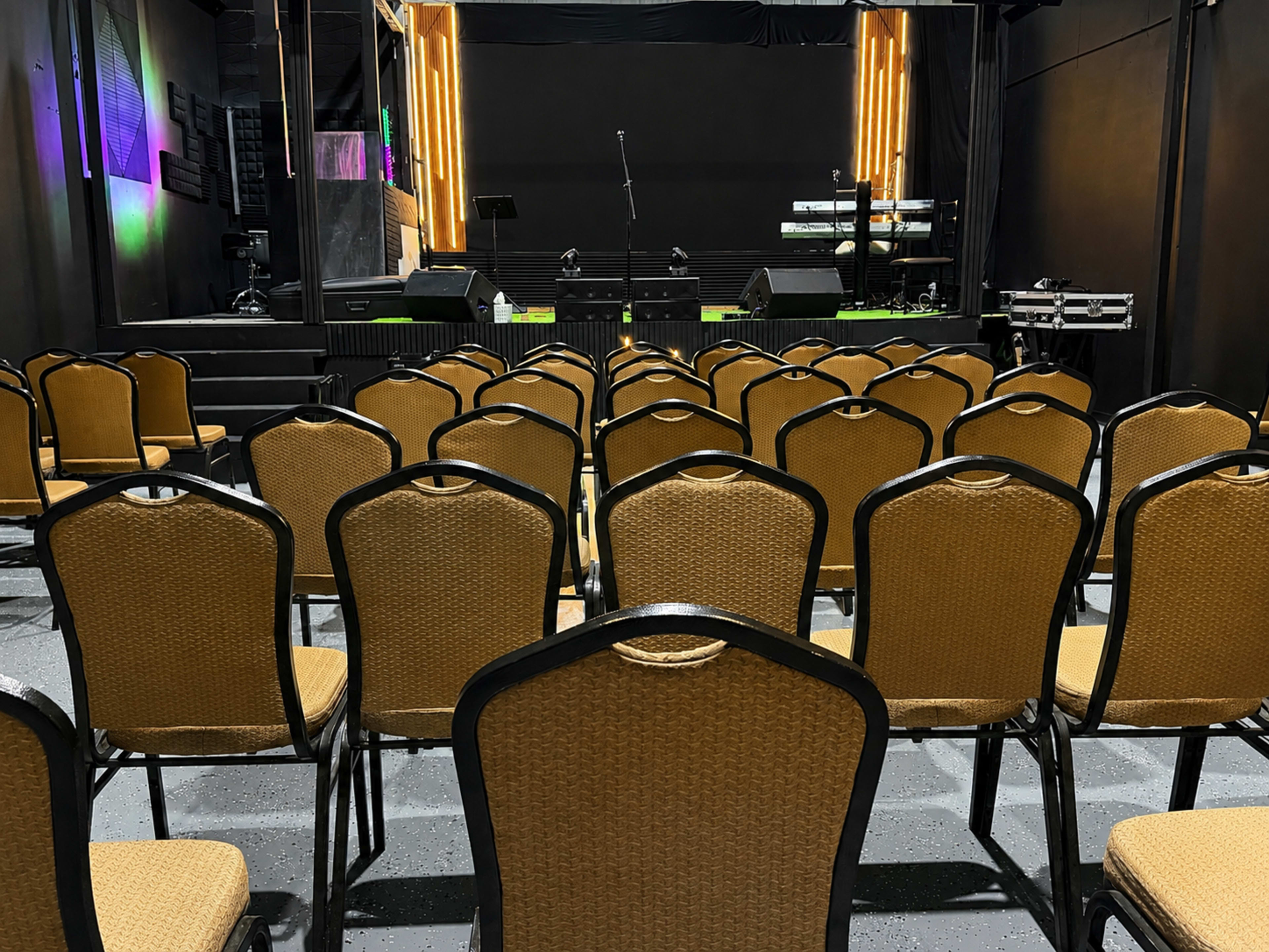 The image shows an empty auditorium with rows of brown chairs facing a stage equipped with musical instruments and lighting.