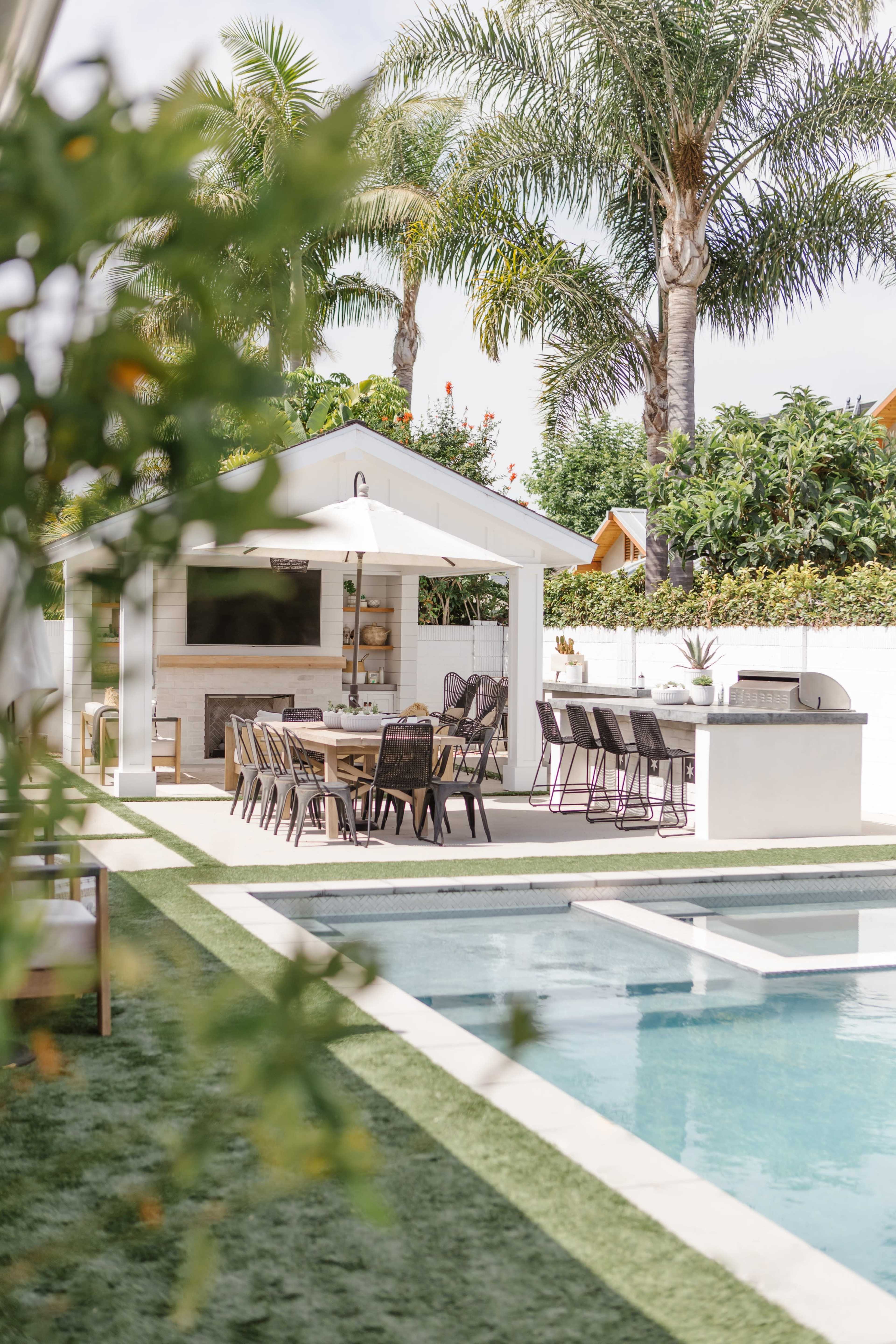 A poolside area features a dining set and a barbecue station surrounded by greenery and palm trees.