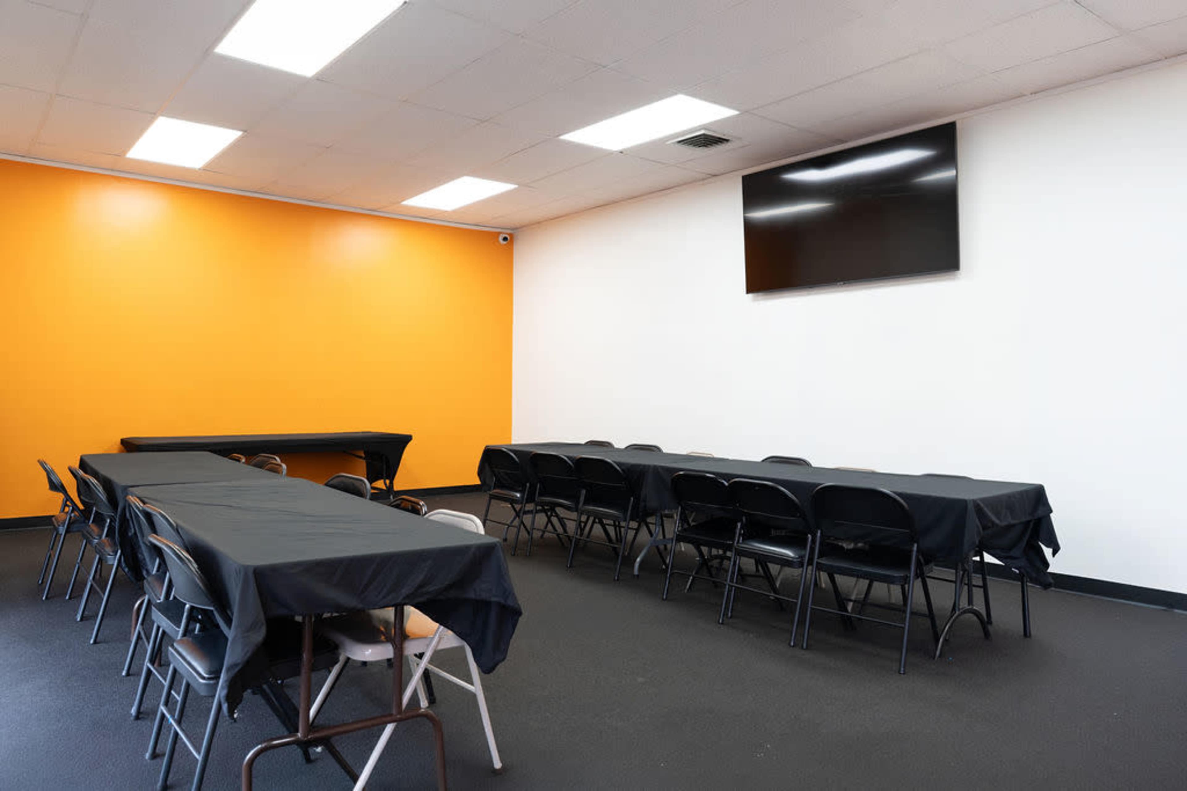 The image shows a meeting room with several rows of black tables and chairs against an orange and white wall, featuring a television mounted on one side.