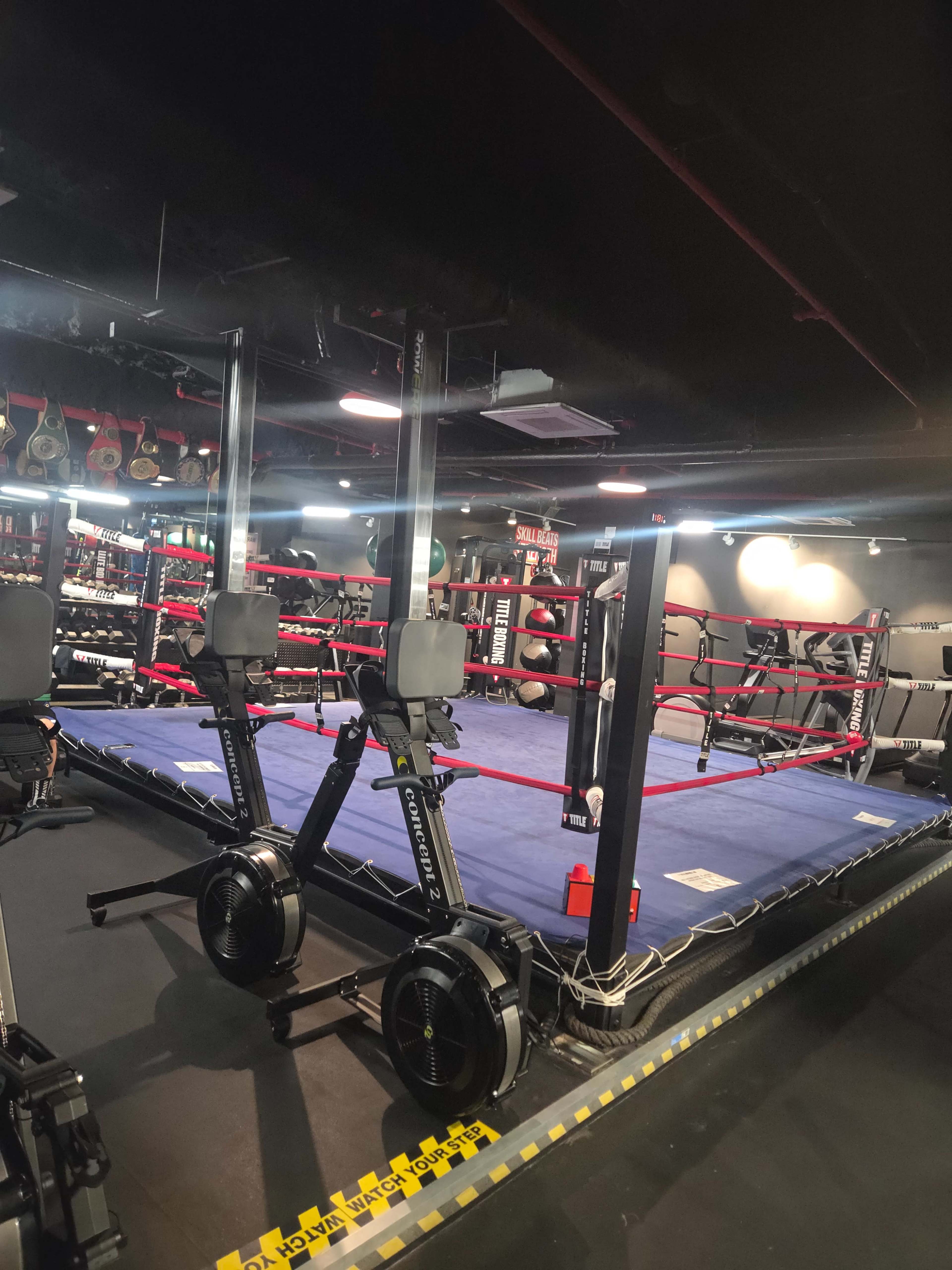 The image shows an indoor boxing gym with a boxing ring in the center and rowing machines positioned in front of it.