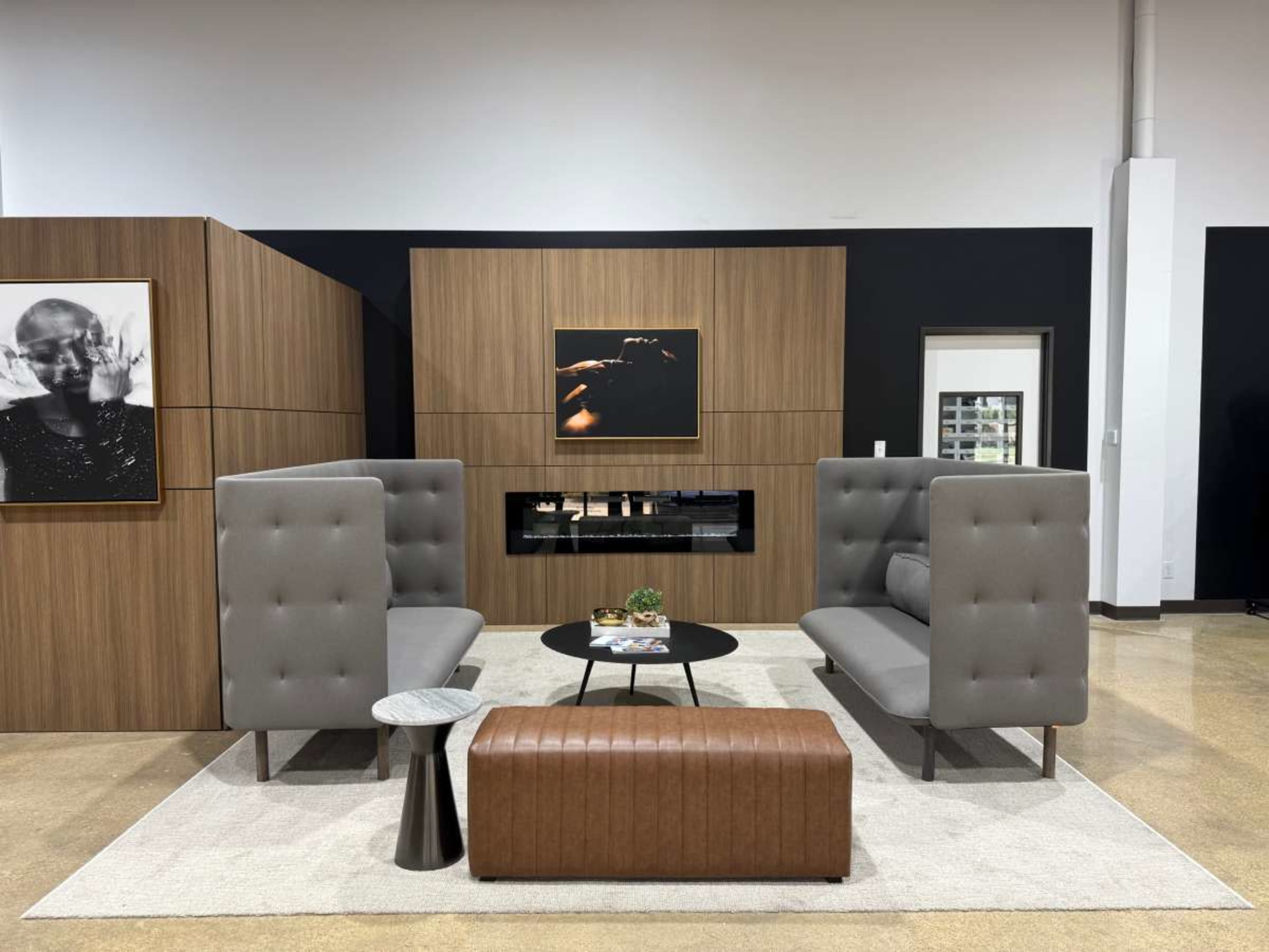 The image shows a modern lounge area featuring two gray upholstered sofas separated by a low black table, with a brown leather bench and framed artwork on the walls.