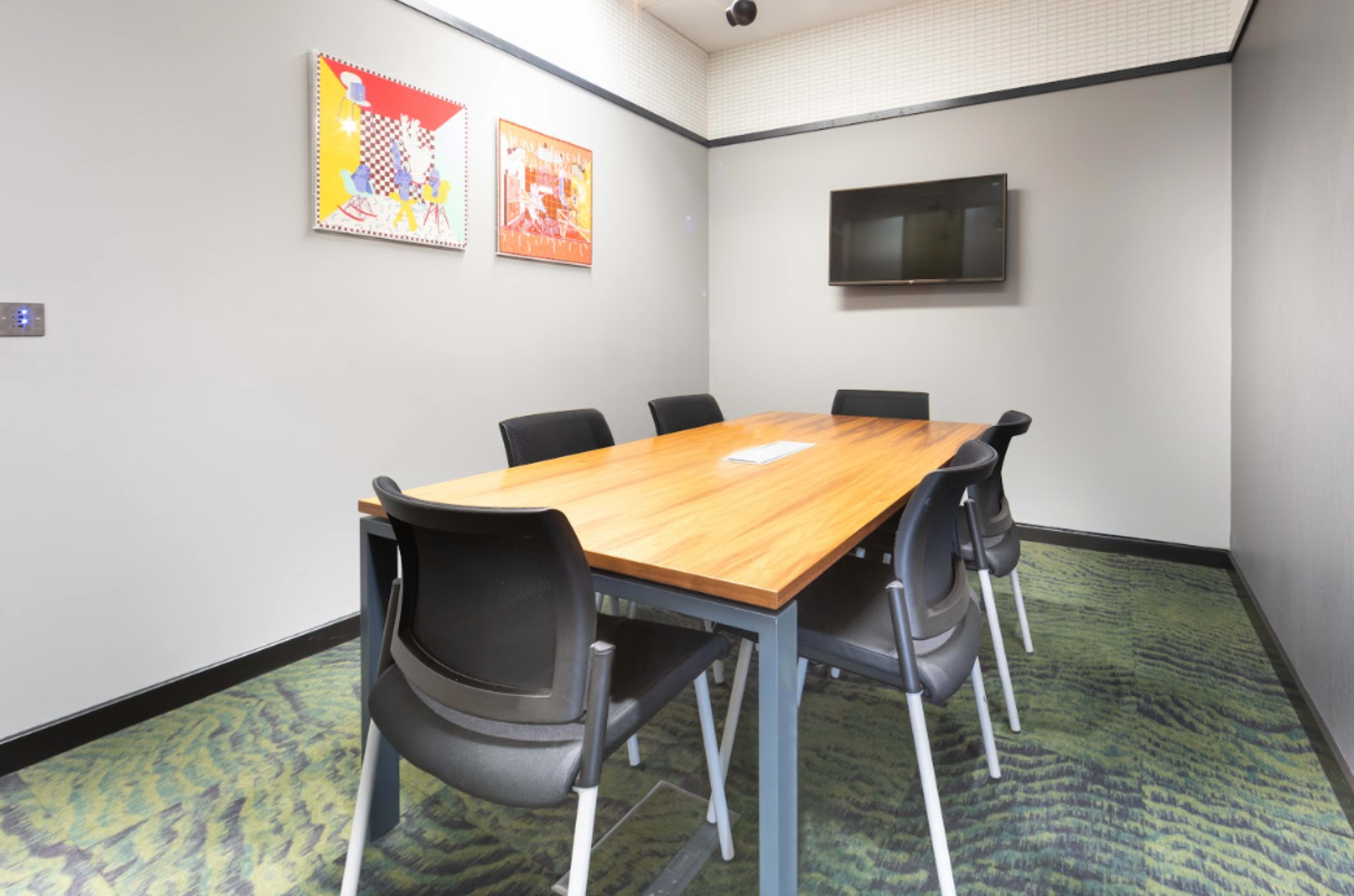 A conference room features a wooden table surrounded by black chairs, with two framed artworks on the wall and a flat-screen TV mounted opposite the table.