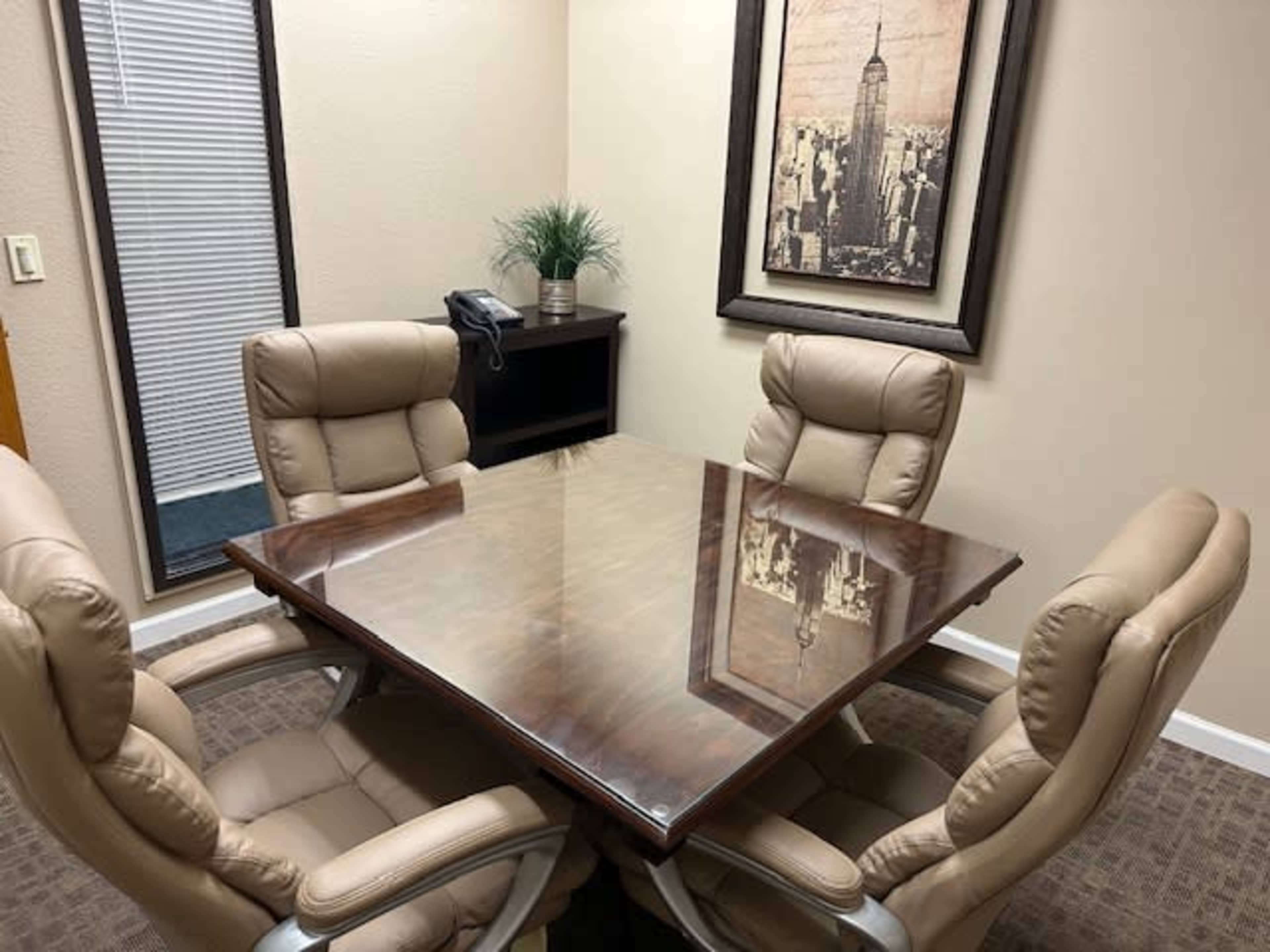 A small conference room with a glass-top table surrounded by four beige leather chairs, and a framed cityscape artwork on the wall.