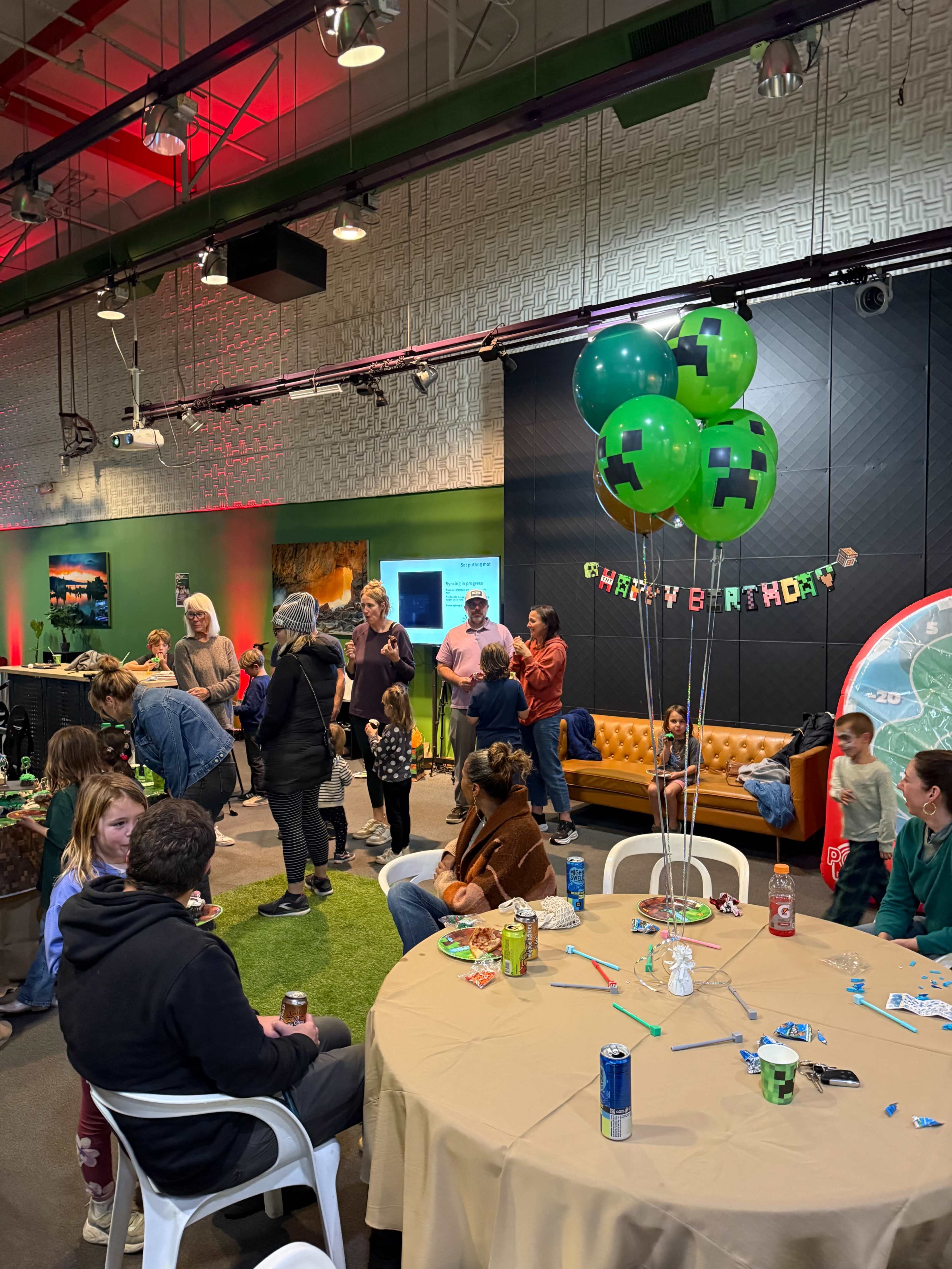 A group of people gathers at a decorated indoor space for a birthday celebration featuring themed balloons and a central table with drinks and party supplies.