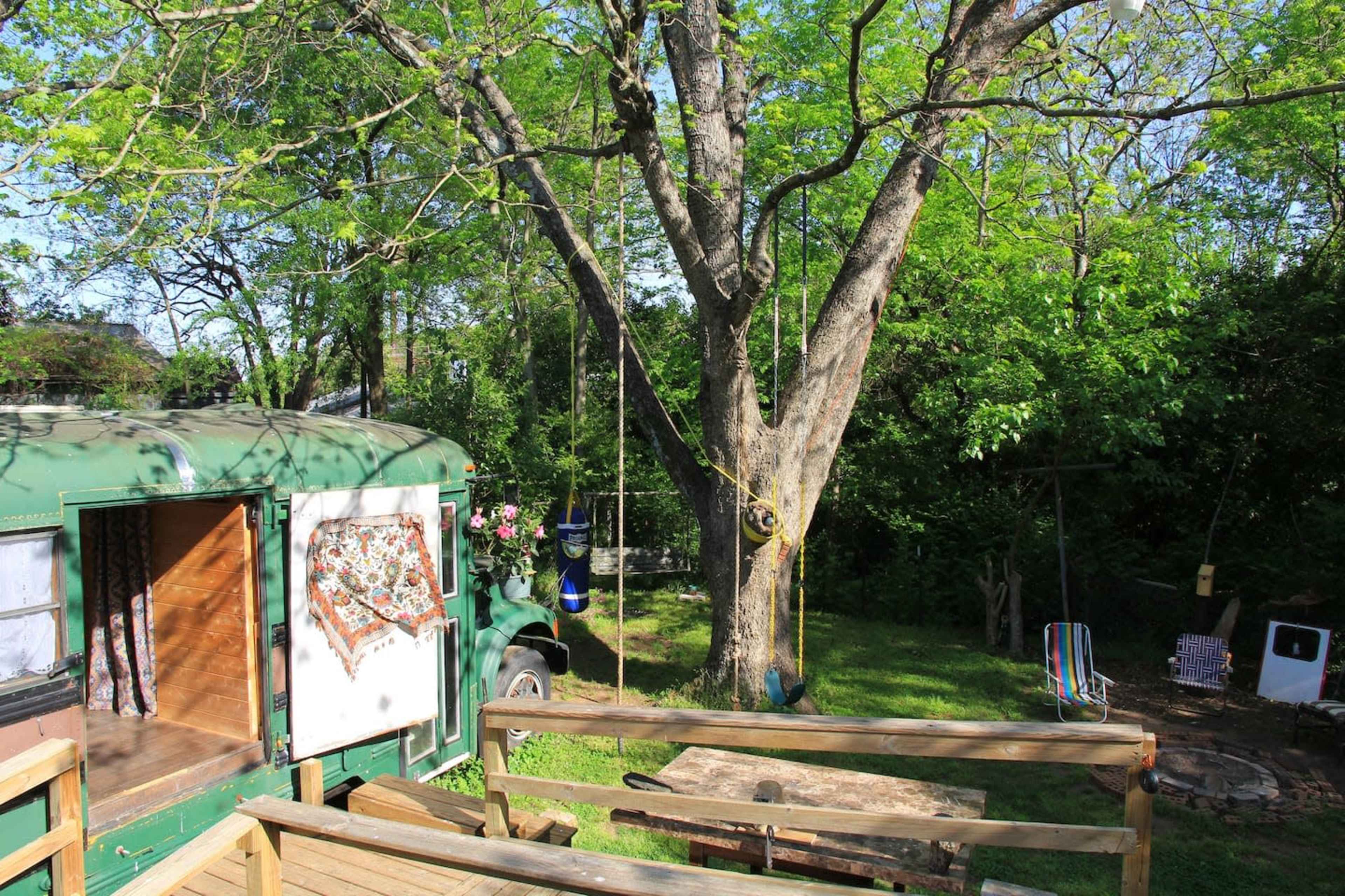 A green bus is parked beside a large tree in a grassy area with a wooden deck and a fire pit nearby.