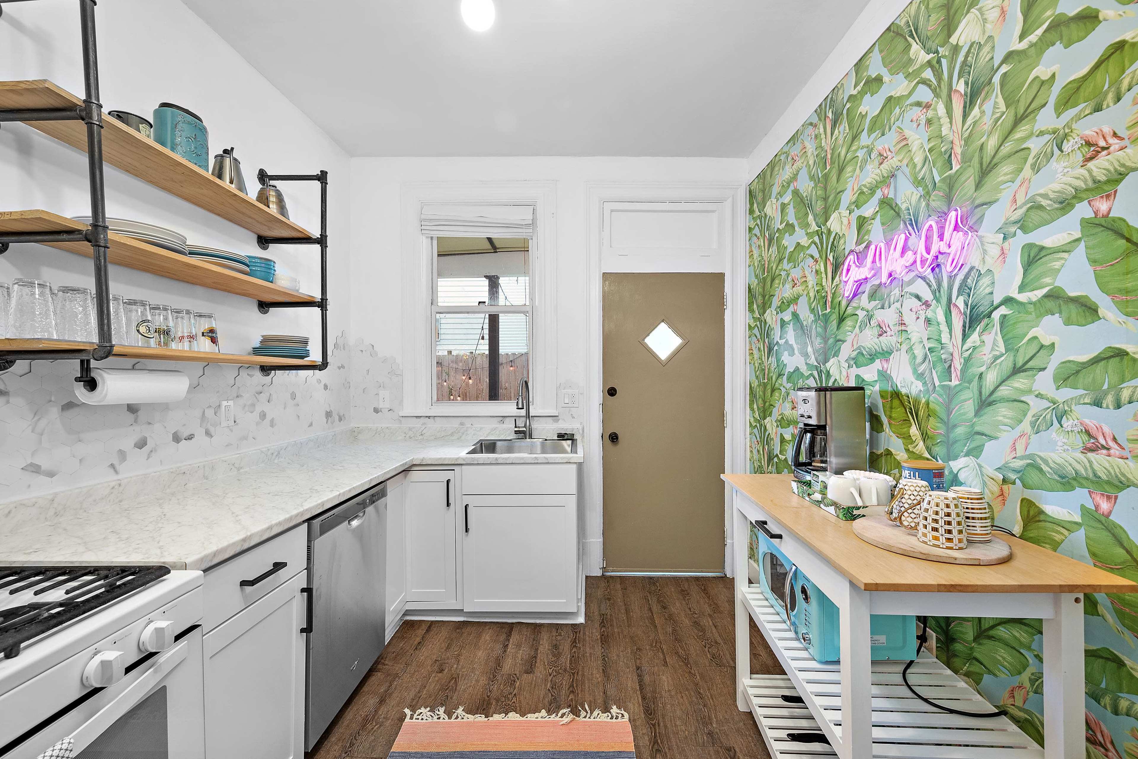 The kitchen features a marble countertop, white cabinets, open shelving with various kitchen items, and a vibrant tropical-themed wallpaper.