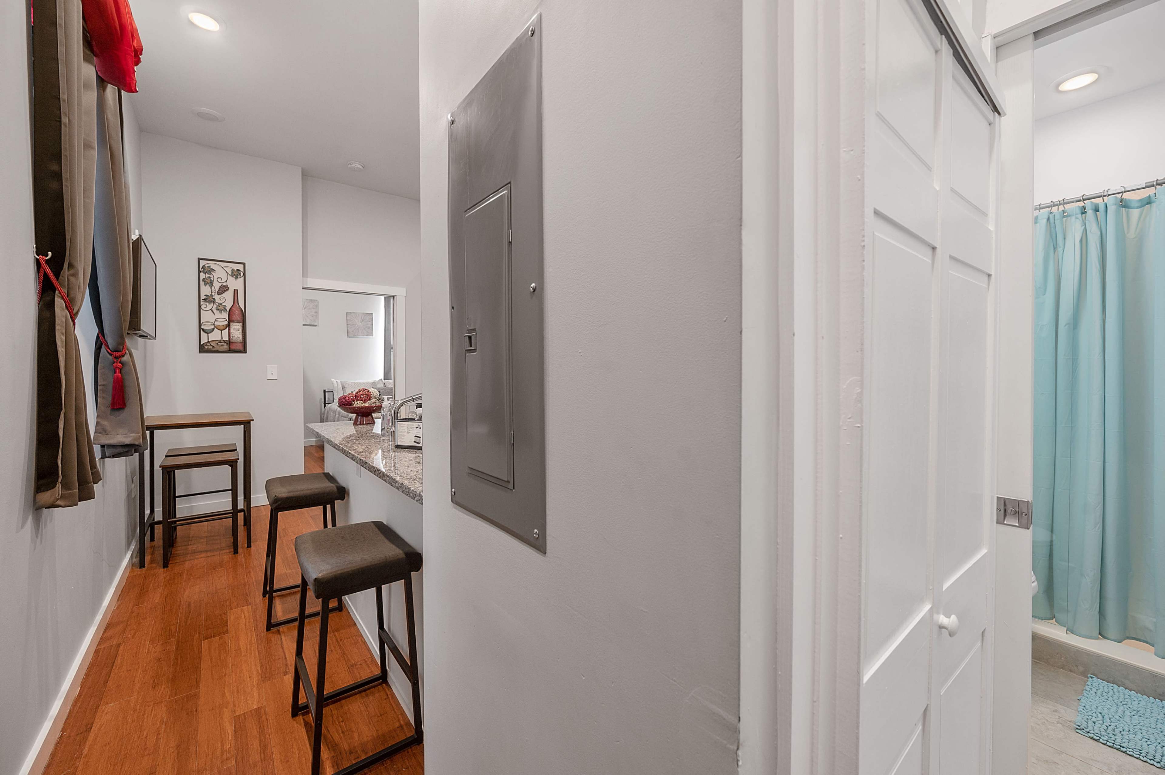A hallway leading to a kitchen area with a countertop and bar stools, a wall-mounted electrical panel, and a bathroom door with a shower curtain visible.