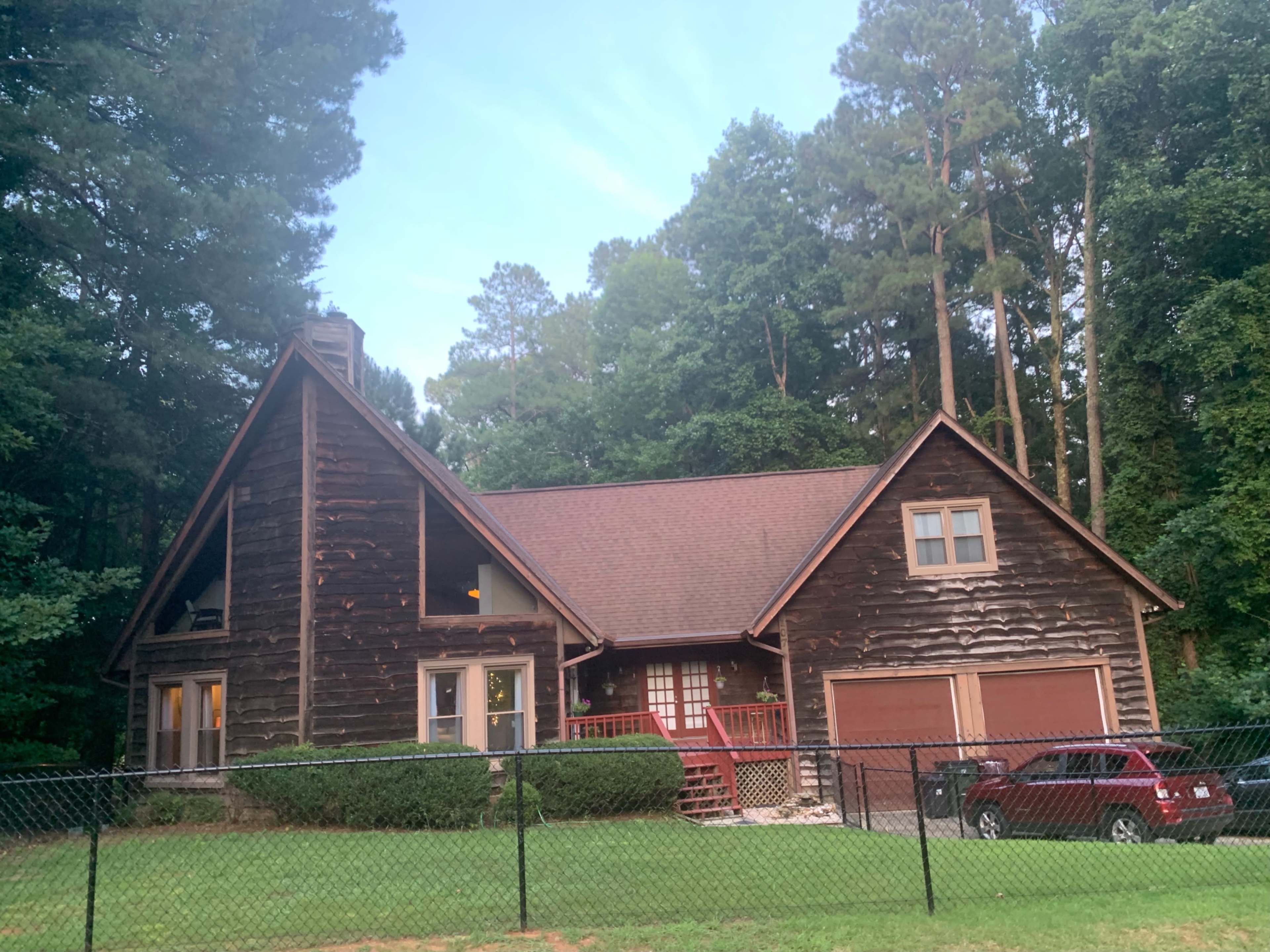A log cabin-style house is surrounded by trees and a fence, with a red vehicle parked in front.