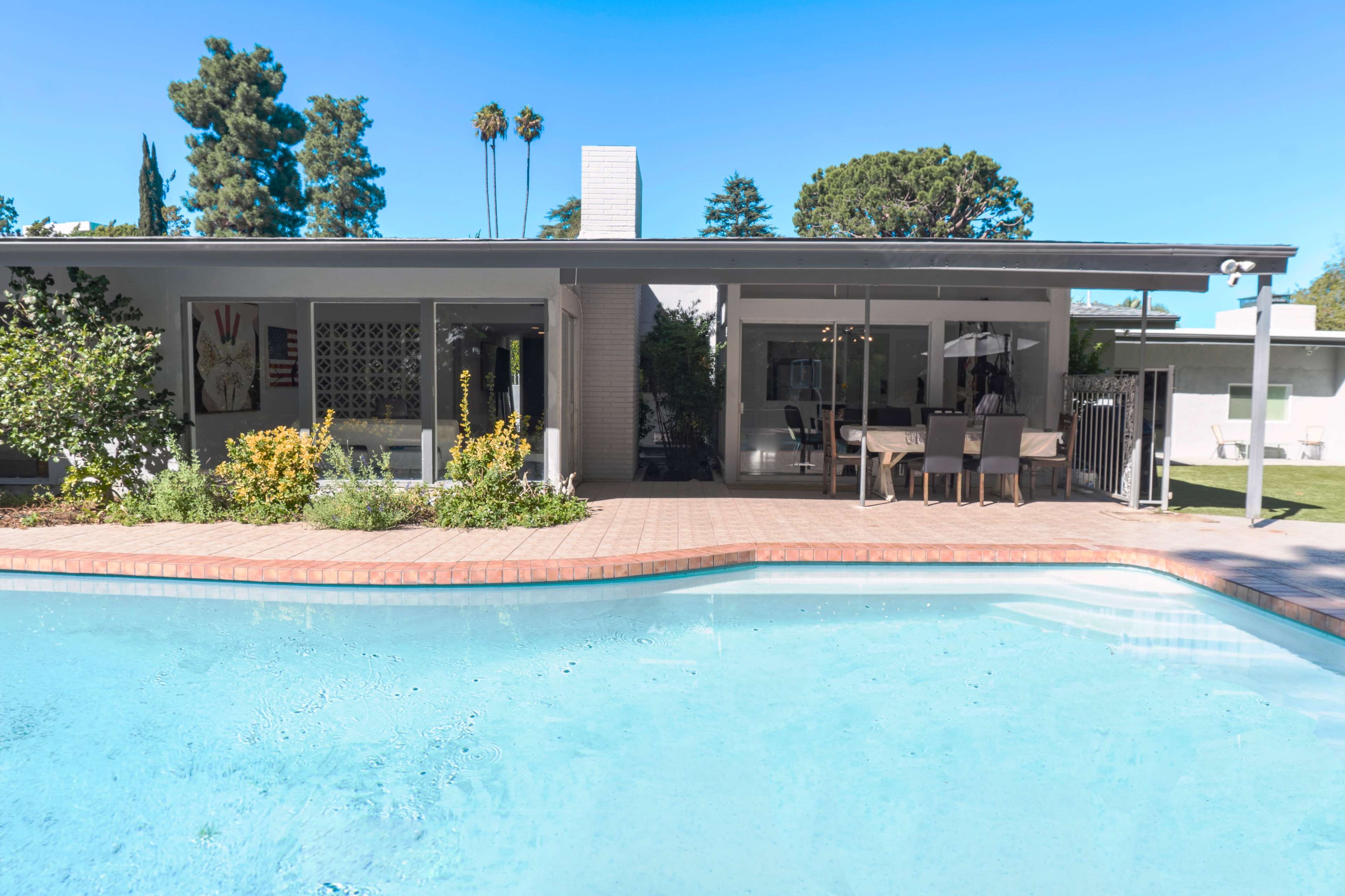 A modern home with a swimming pool and outdoor dining area is surrounded by greenery and trees.