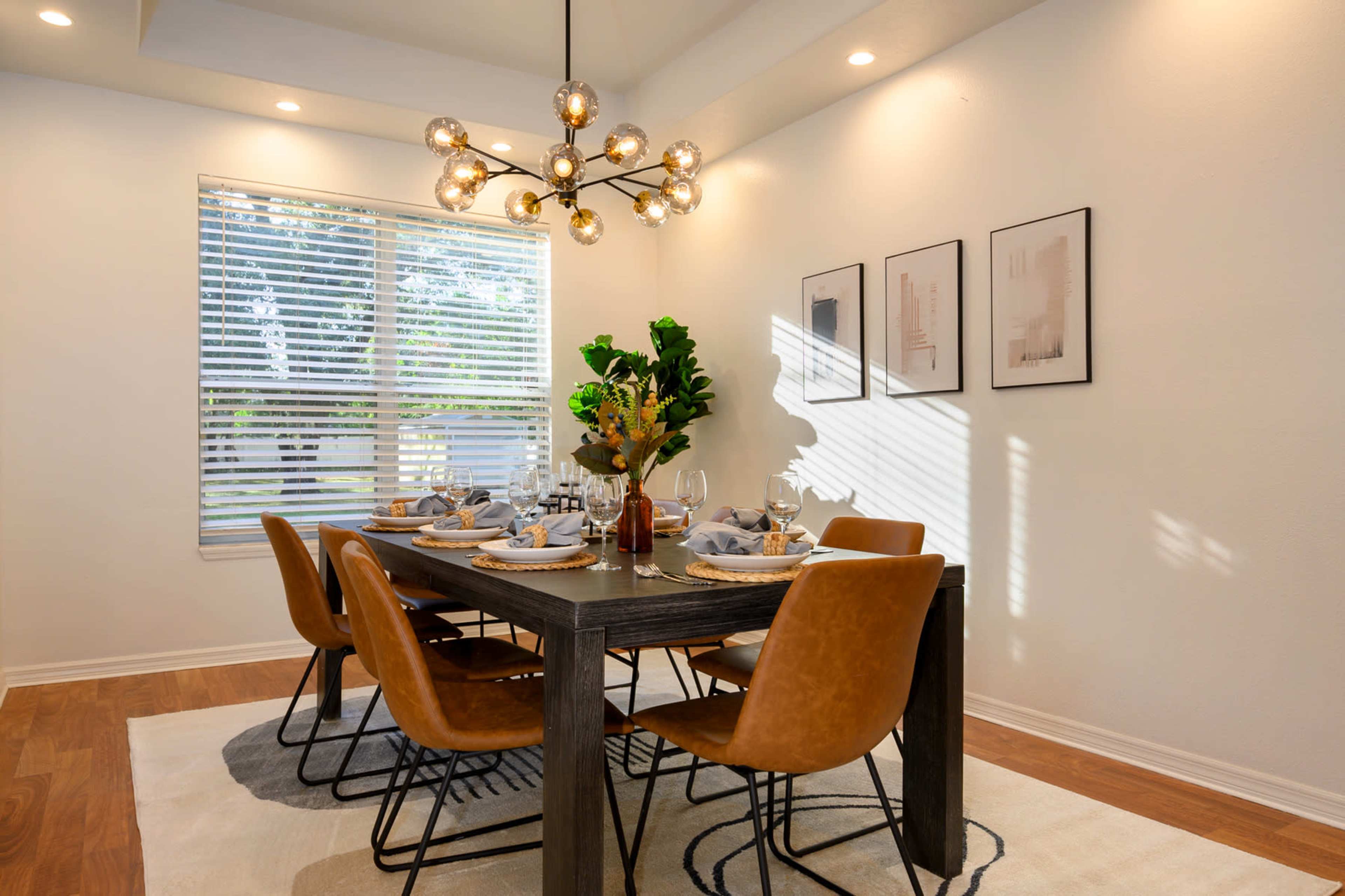 A modern dining room features a table set for six with brown chairs, a decorative centerpiece, and framed artwork on the walls, illuminated by a stylish light fixture.