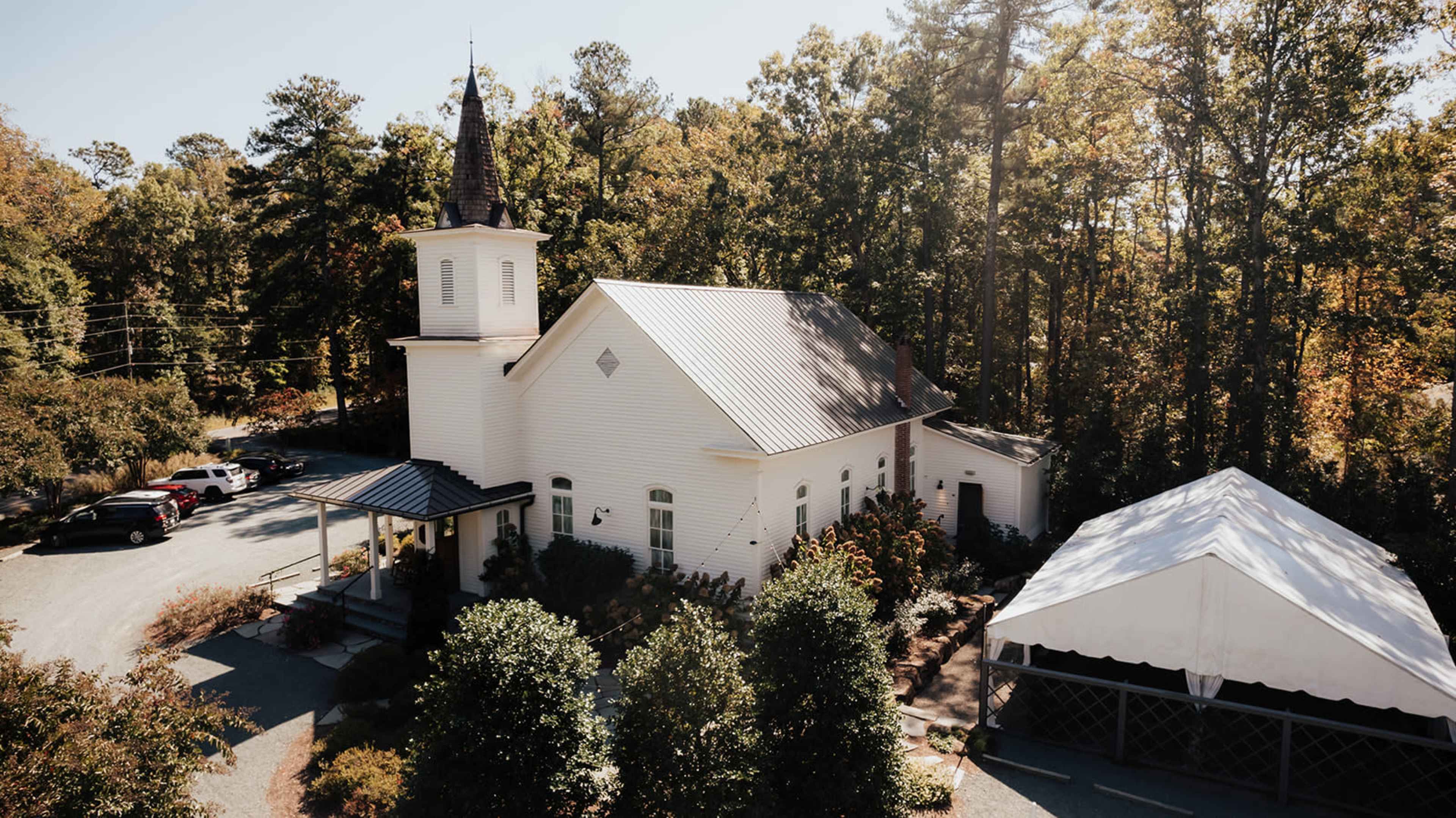 A white church with a steeple is nestled among trees, adjacent to a large tent and a gravel parking area.