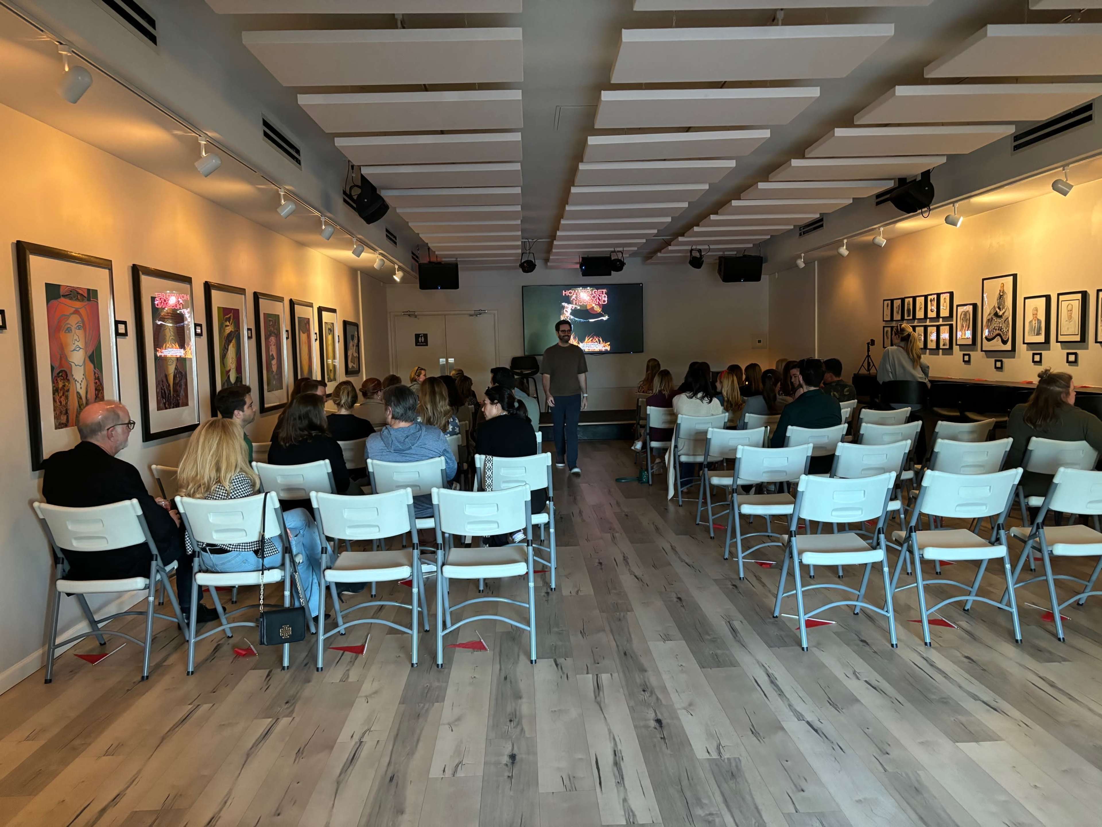 A group of people sits in rows of chairs facing a presenter in a gallery-like space with framed artworks on the walls.